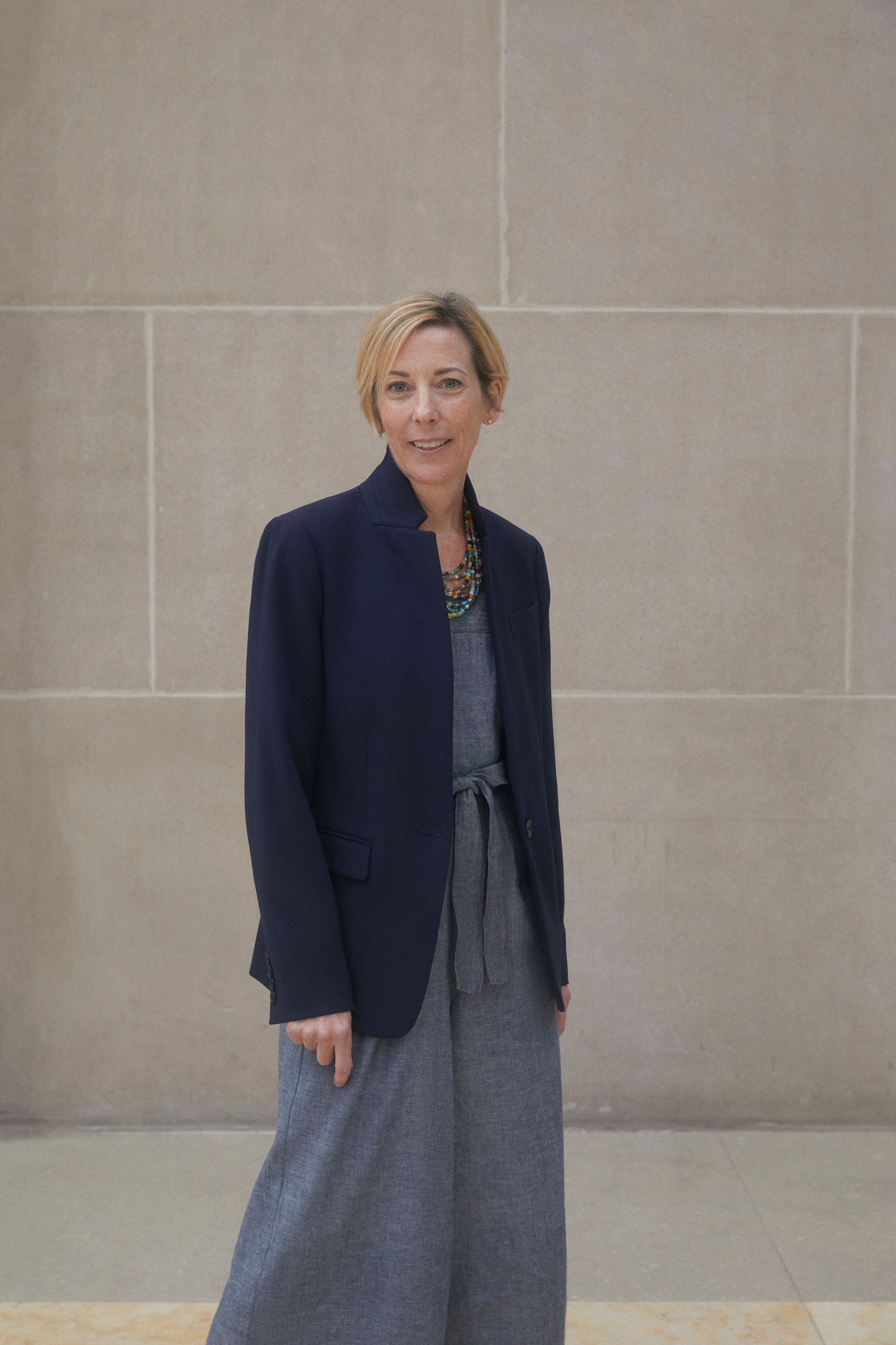 A color photograph of Kerry Mullins standing in front of a stone wall in a long light blue dress and blue blazer.