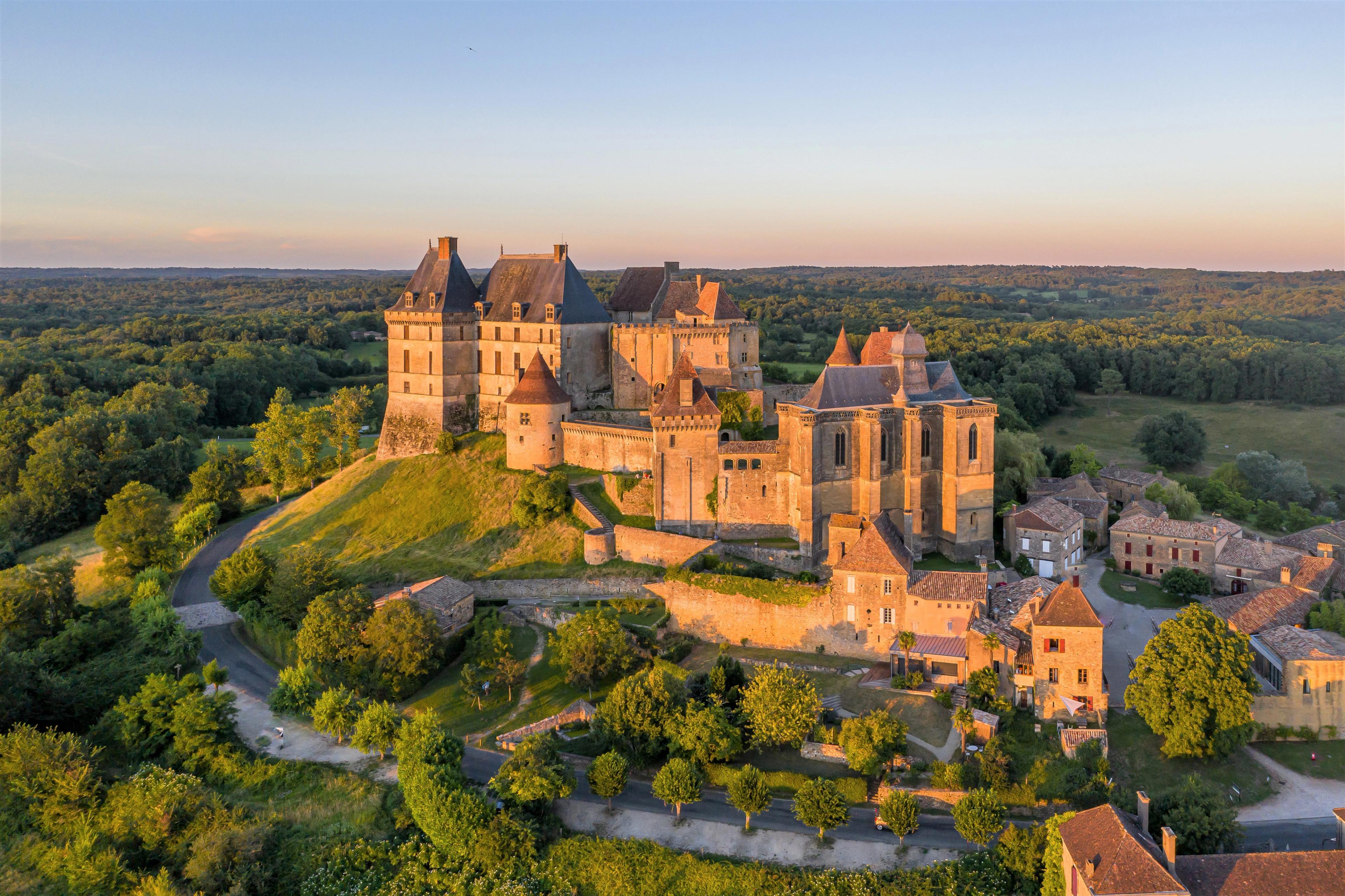 Picturesque castle with numerous pointed roofs and turrets, situated atop a hill and surrounded by green trees.