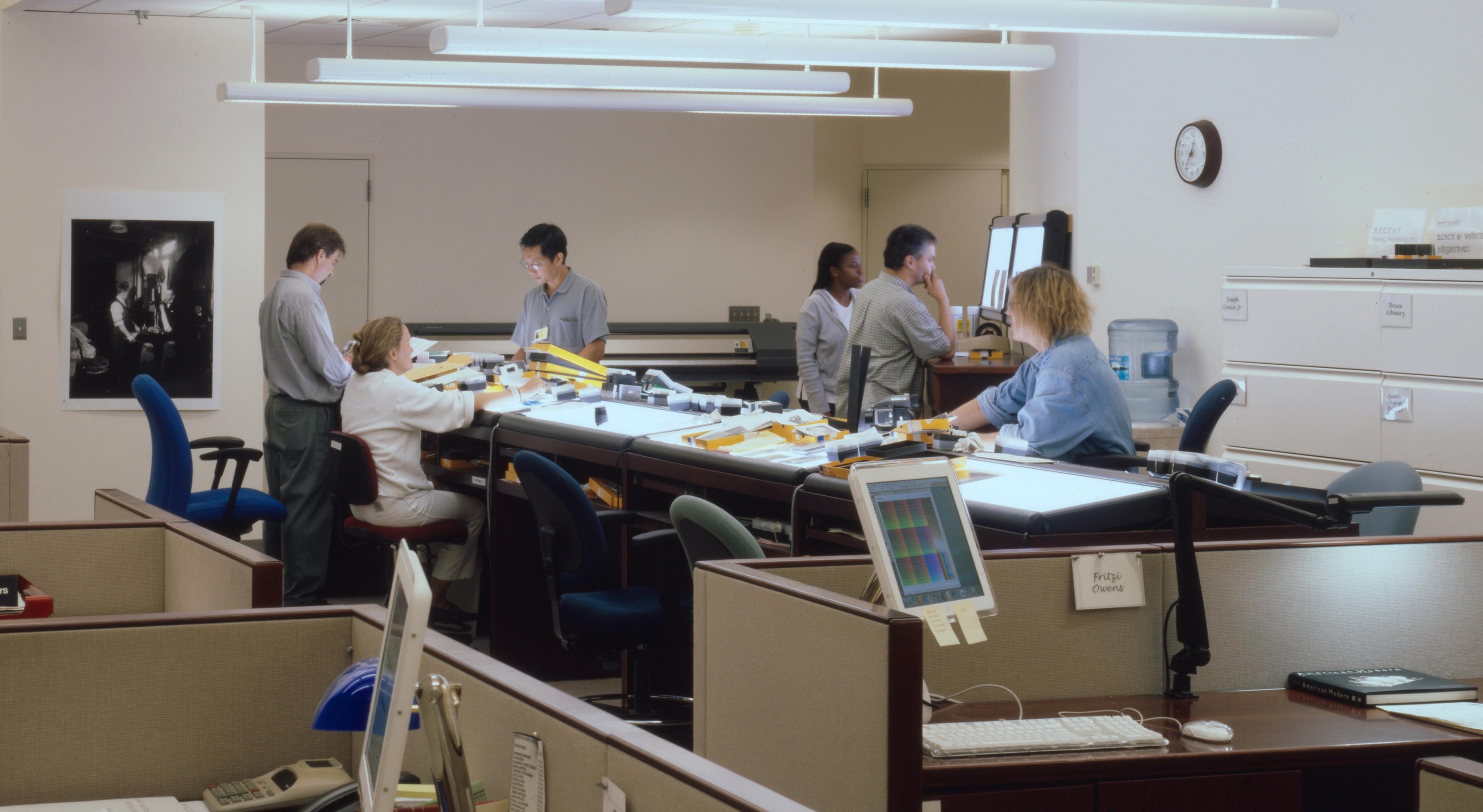 Several people work at a long table with both overhead and backlighting in an office setting.