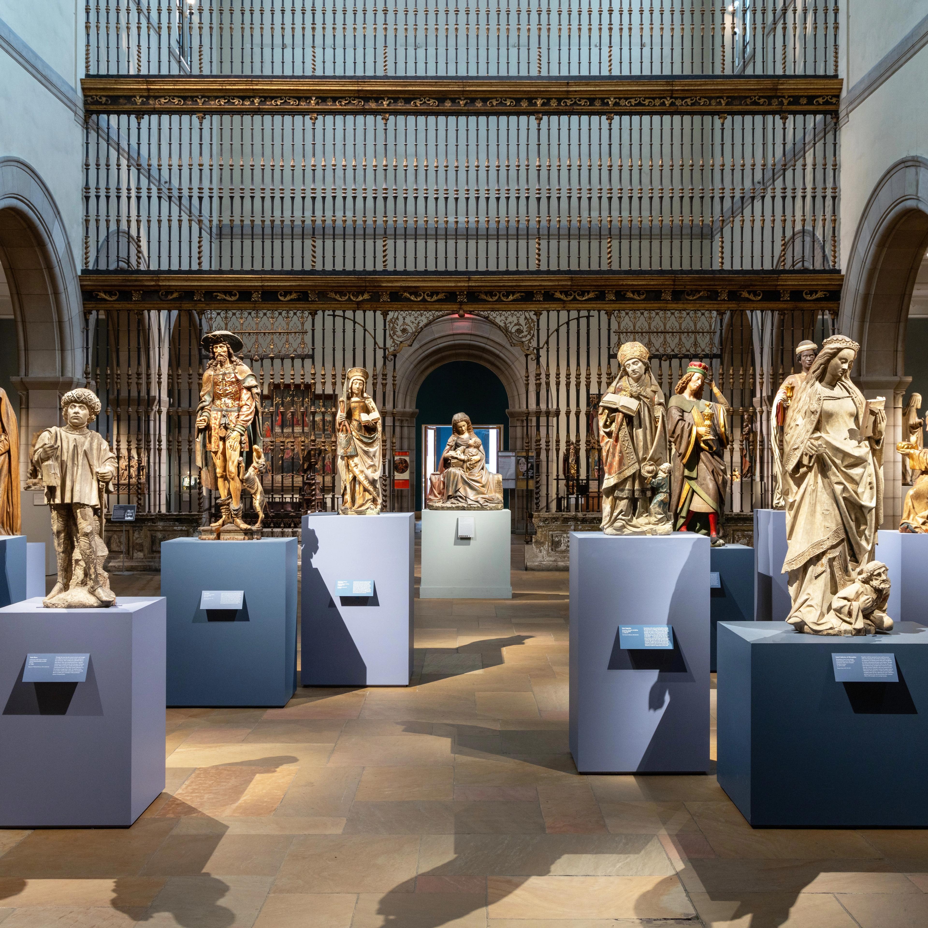 Medieval sculptures displayed on pedestals in a softly lit gallery space with stone arches and a Spanish choir screen.