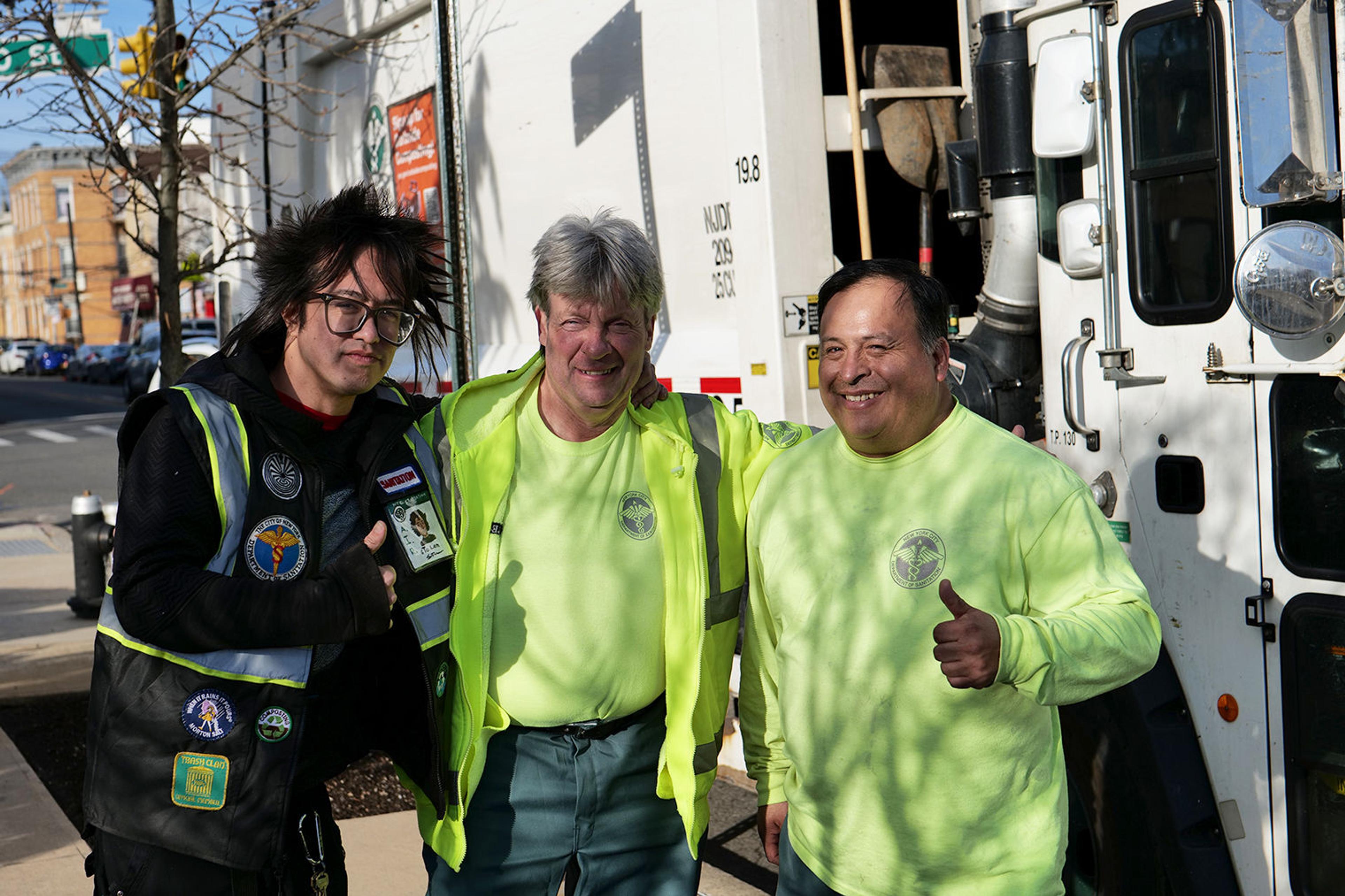 Photograph of three people in black and bright green Sanitation Department attire. They are posing outside in front of a box truck.