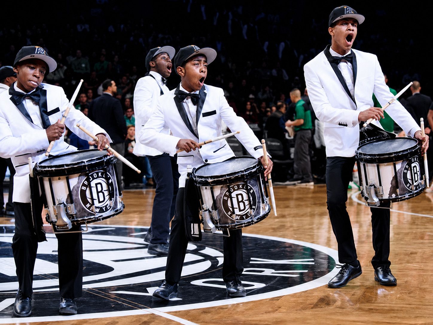 A group of young drummers in white jackets, bowties, and baseball caps play on a basketball court.