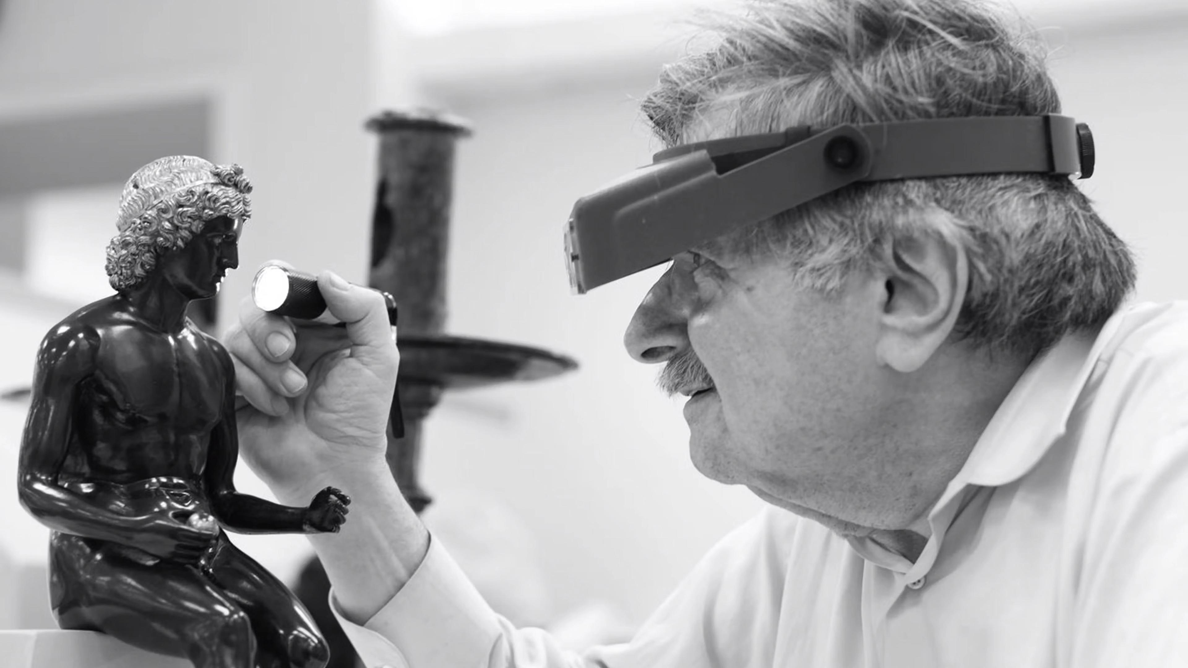 Black and white photograph of a man holding a flashlight and looking closely at a small sculpture.