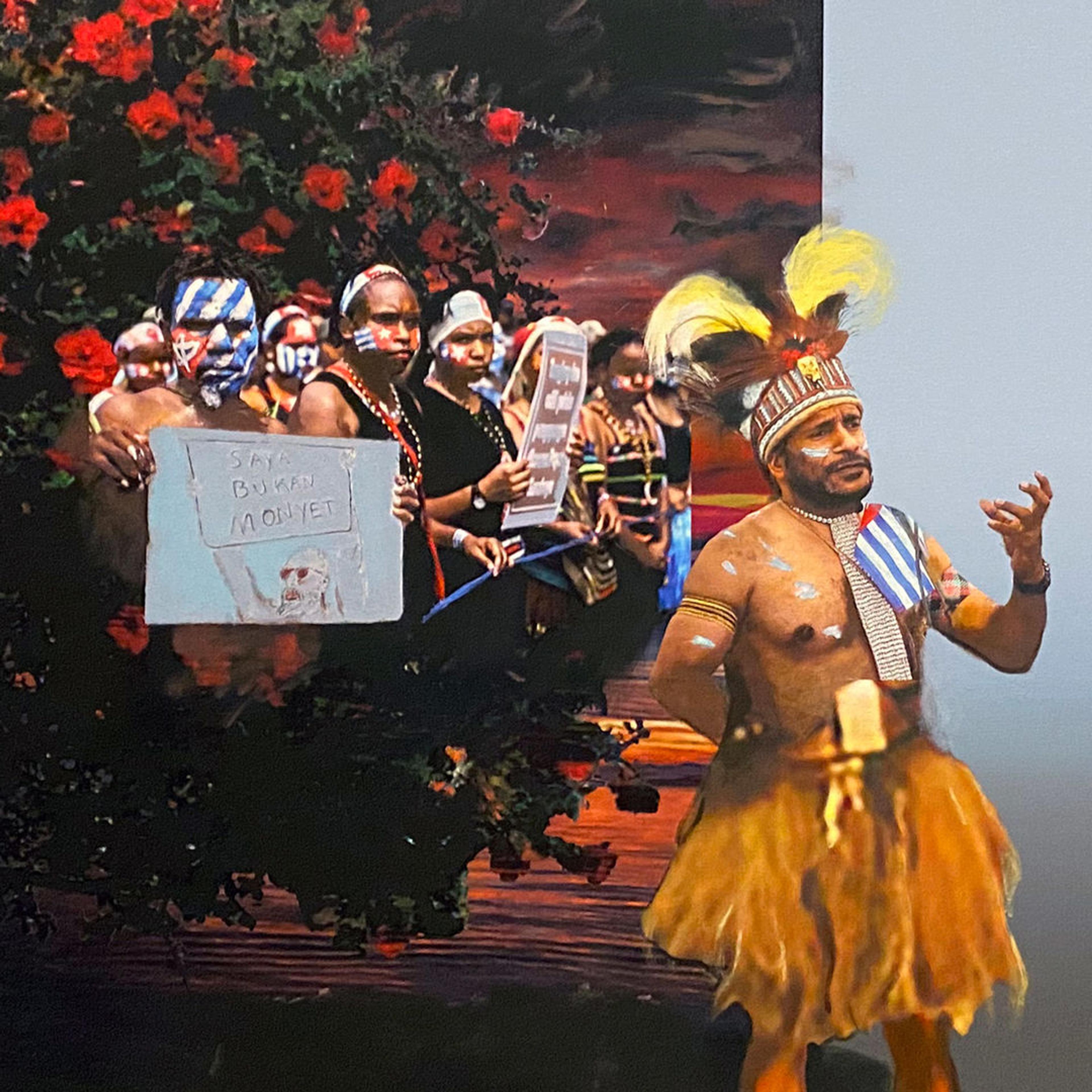 A crowd of people to the left hold up a sign while a shirtless man holds his left hand up as in an oratory gesture