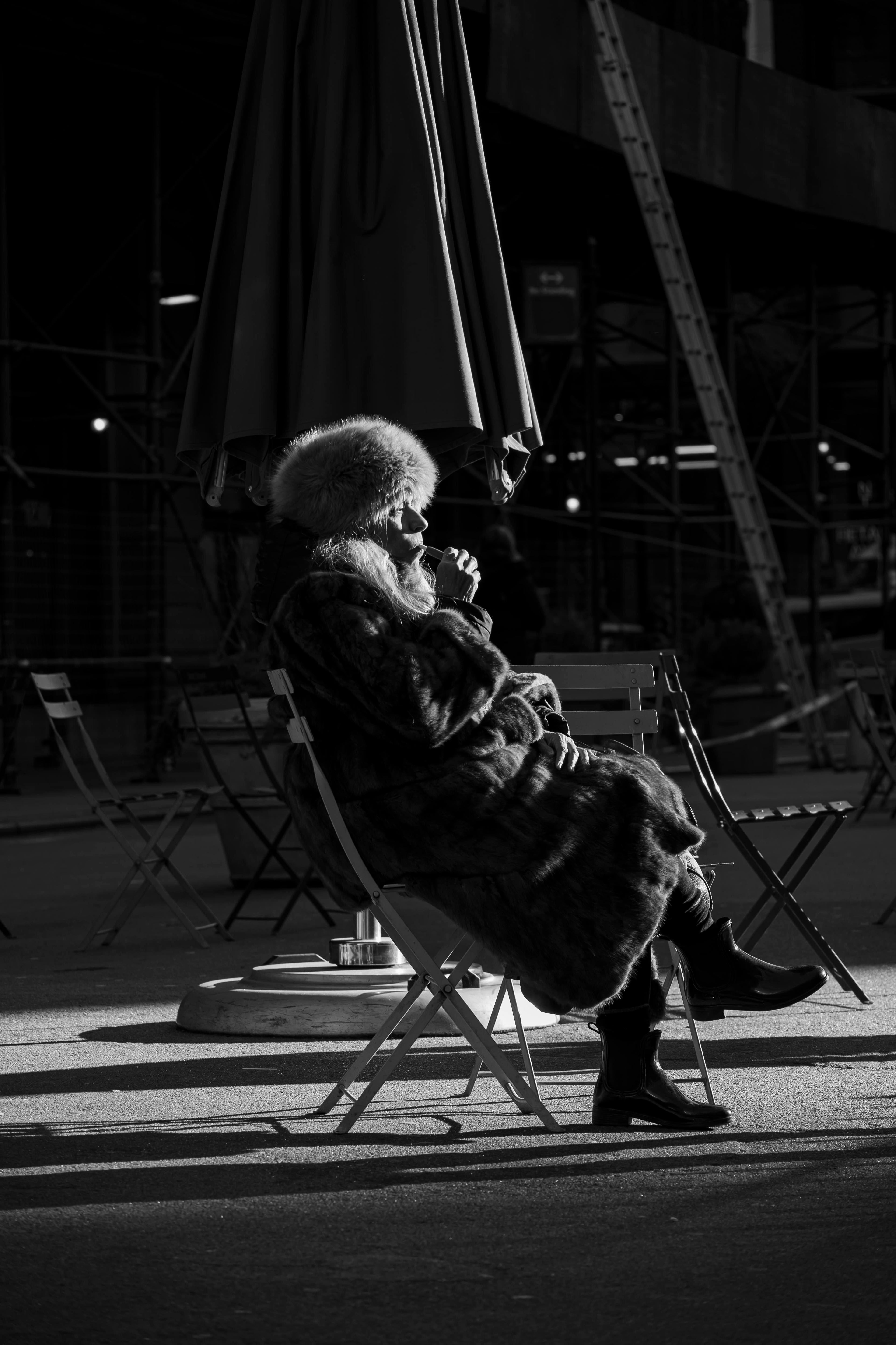 Black-and-white digital photograph of a woman sitting outdoors at night on a wooden folding chair, facing to the right, cast in extreme shadow with contrasting light that illuminates her from the right. Her legs are crossed at the knee, and she wears winter boots, a thick winter coat, and a furry Cossack hat. With her right hand, she holds what might be a pen or a pipe to her lips. Other wooden folding chairs and a collapsed awning on a stand appear in the background, along with a tall, fully extended ladder in the pitch-black distance.