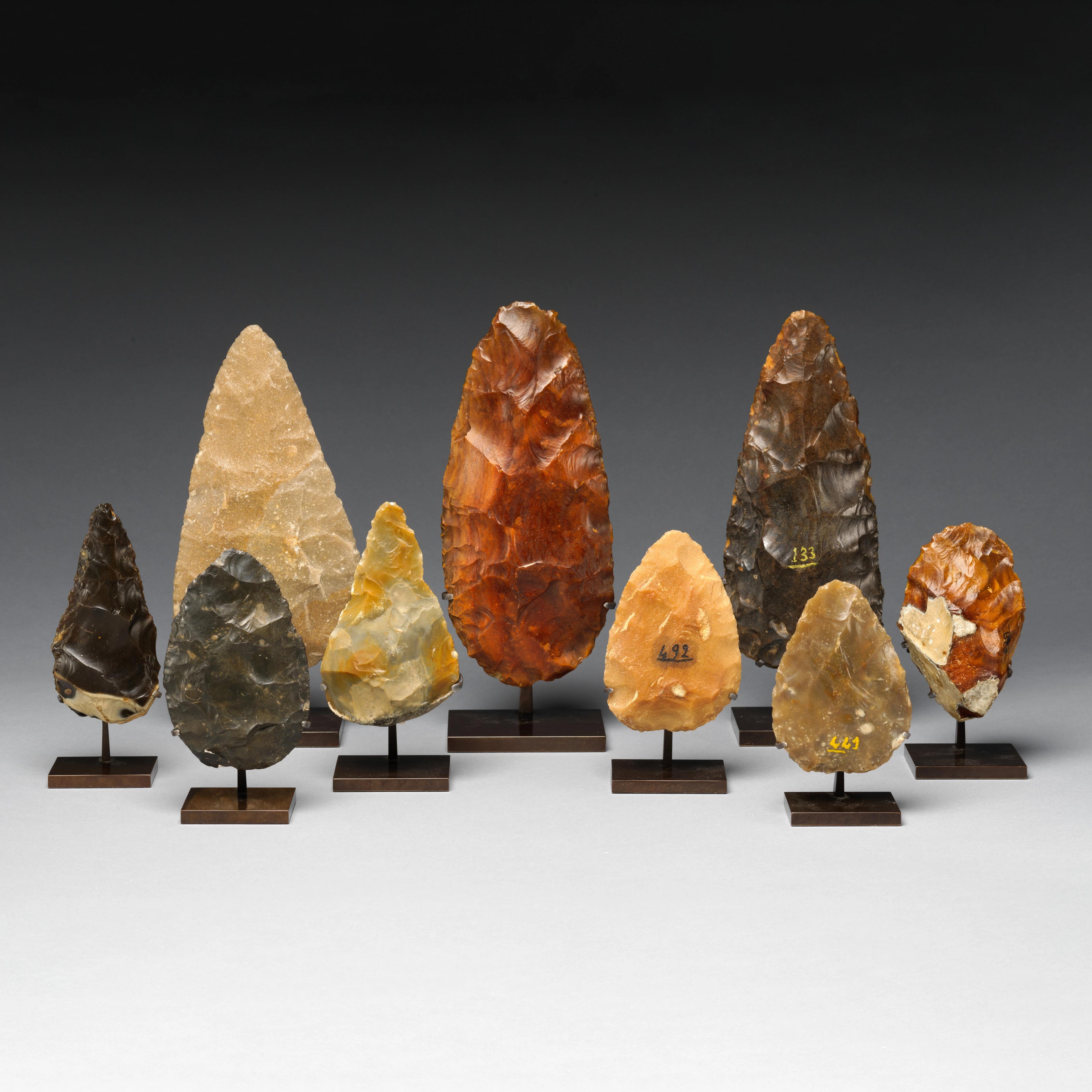 Set of nine bifaces, carved delicately into spears, placed upright against a gray background.