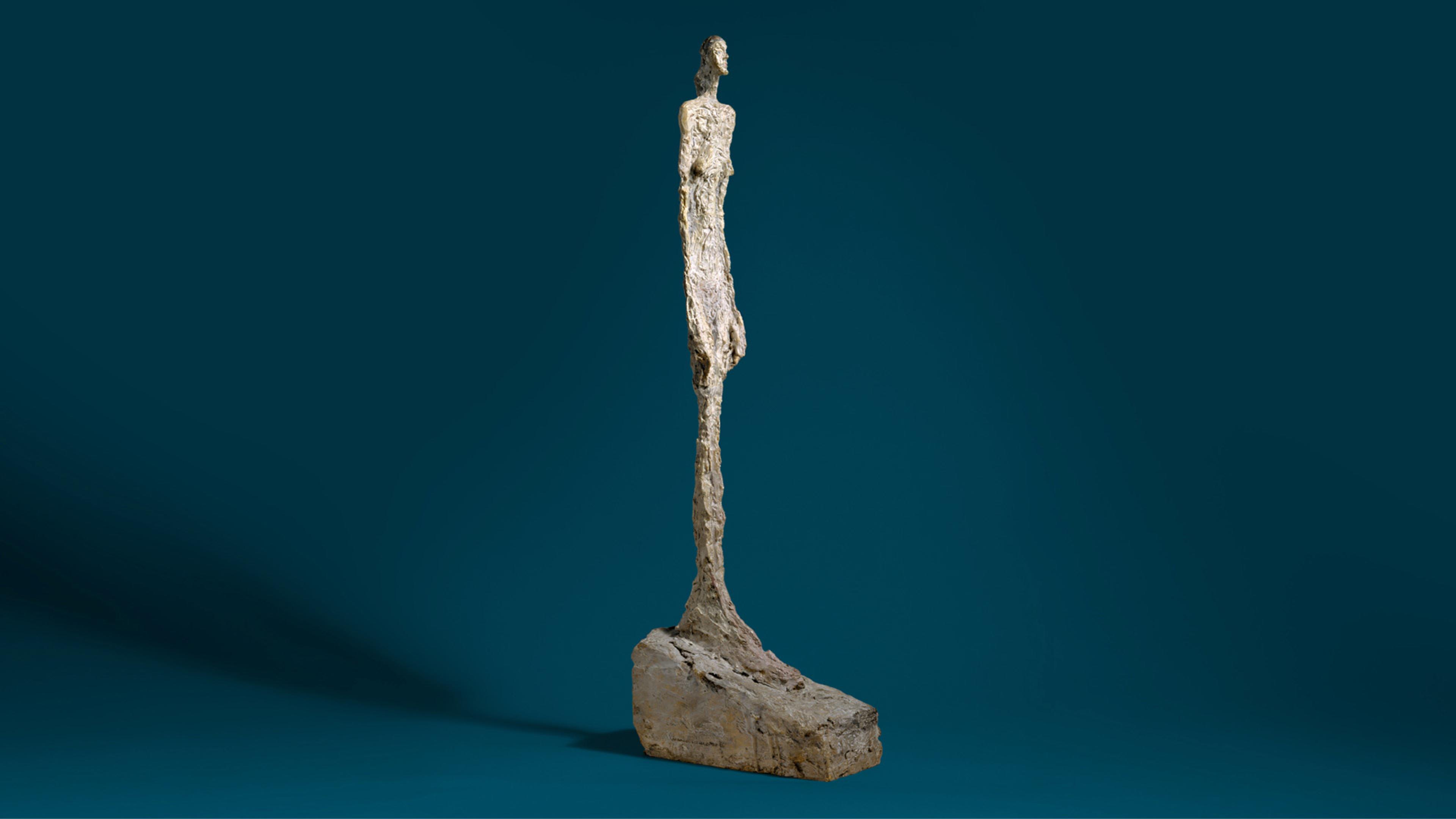 Slender, textured statue of a figure with elongated feet and a thick base against a deep blue background.