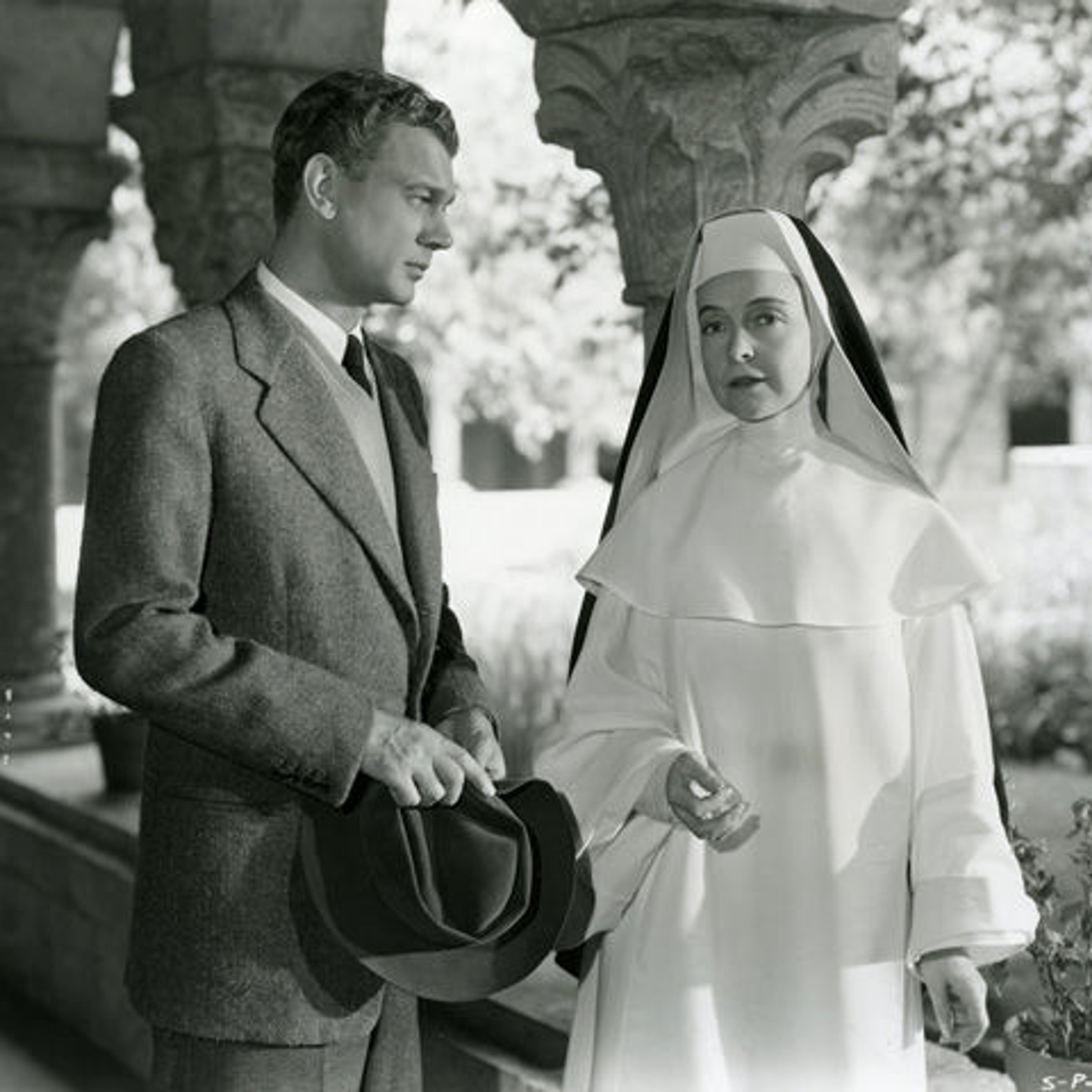 A well-dressed man and a nun wearing white robes stand together in the Cuxa Cloister.