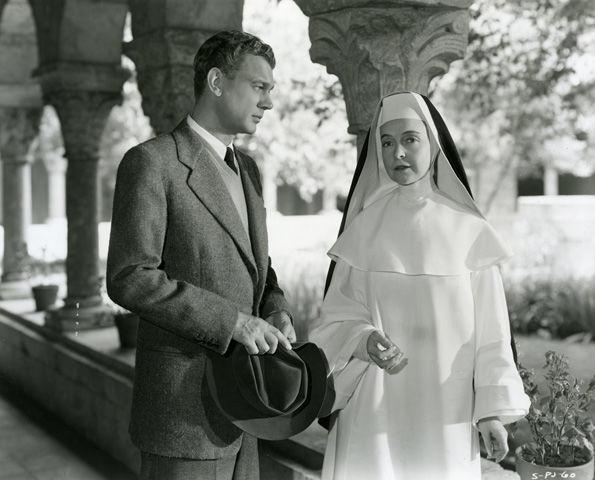 A well-dressed man and a nun wearing white robes stand together in the Cuxa Cloister.