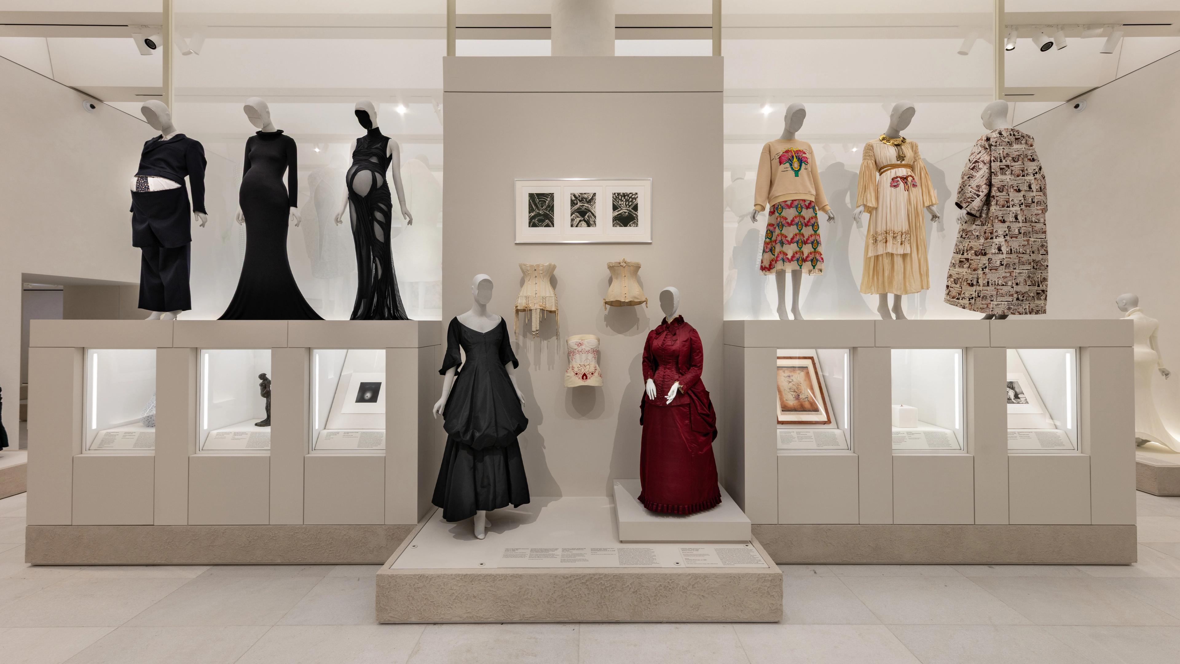 A fashion exhibit showcasing mannequins in diverse historical garments. Outfits range from elegant black dresses to colorful traditional attire
