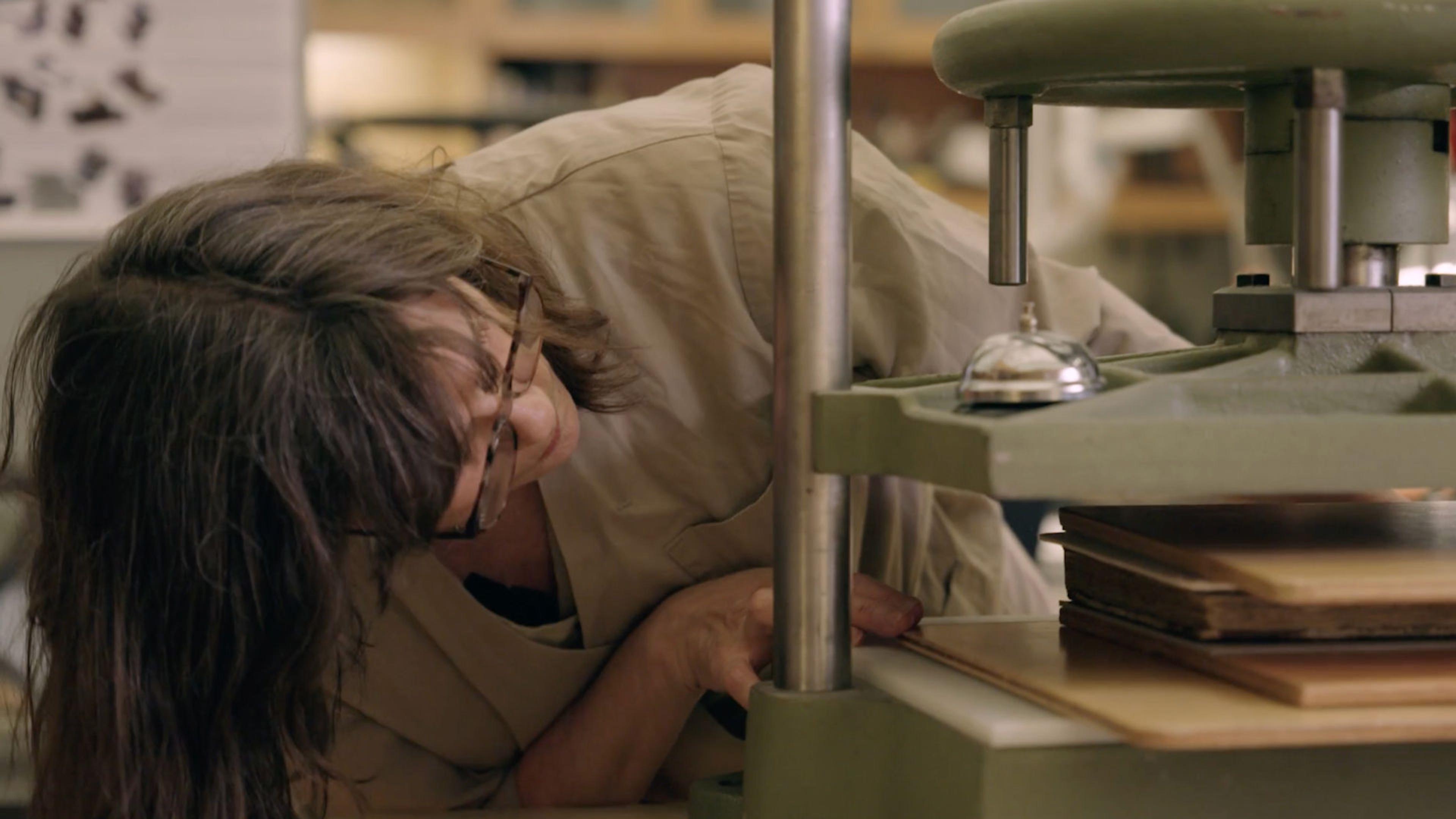 A person leans over to place a book between the plates of a book press in the foreground at right.