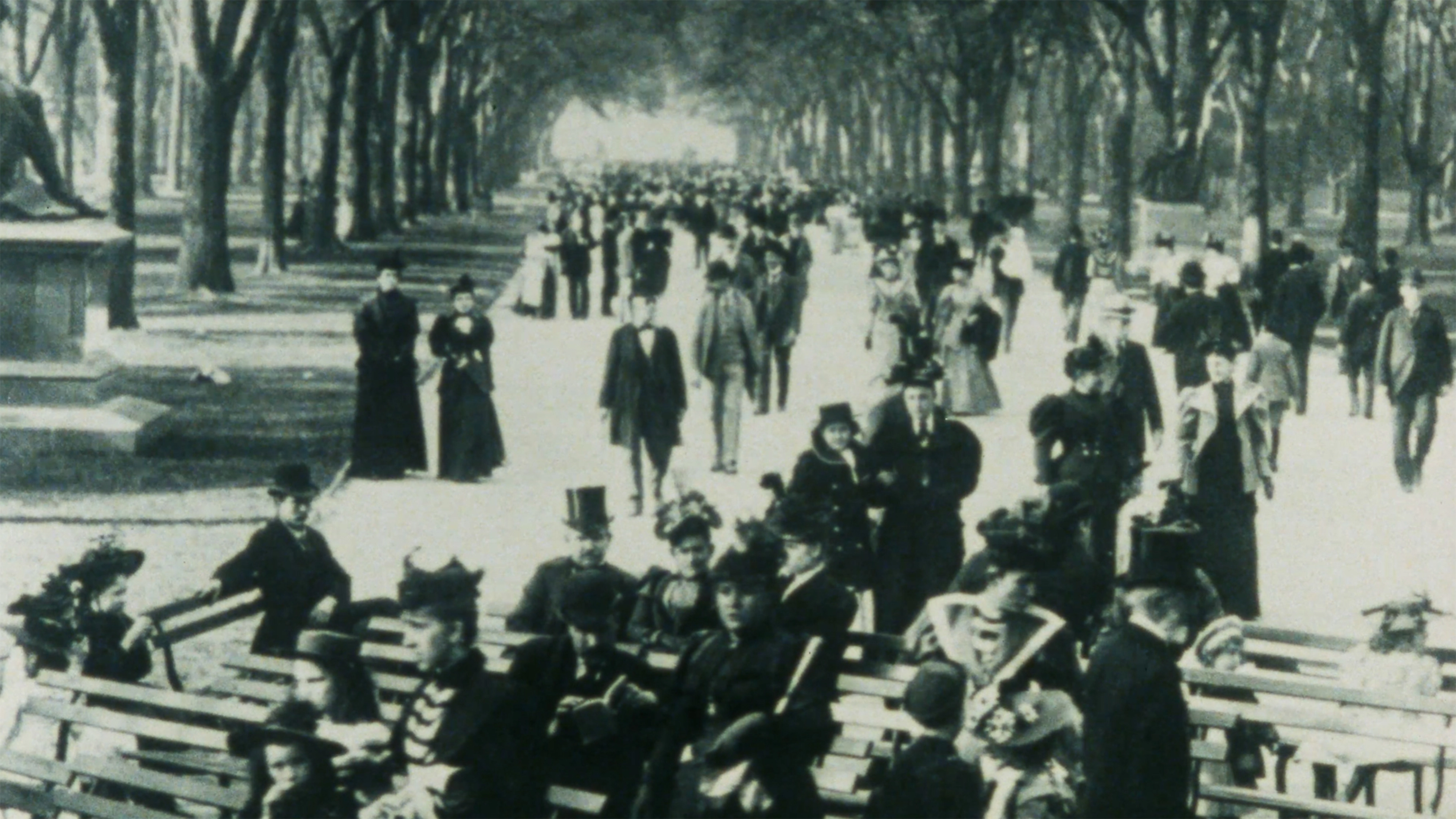 Vintage photo of a busy city park walkway lined with trees. People in formal 1900s attire, including hats and suits, walk and sit on benches.