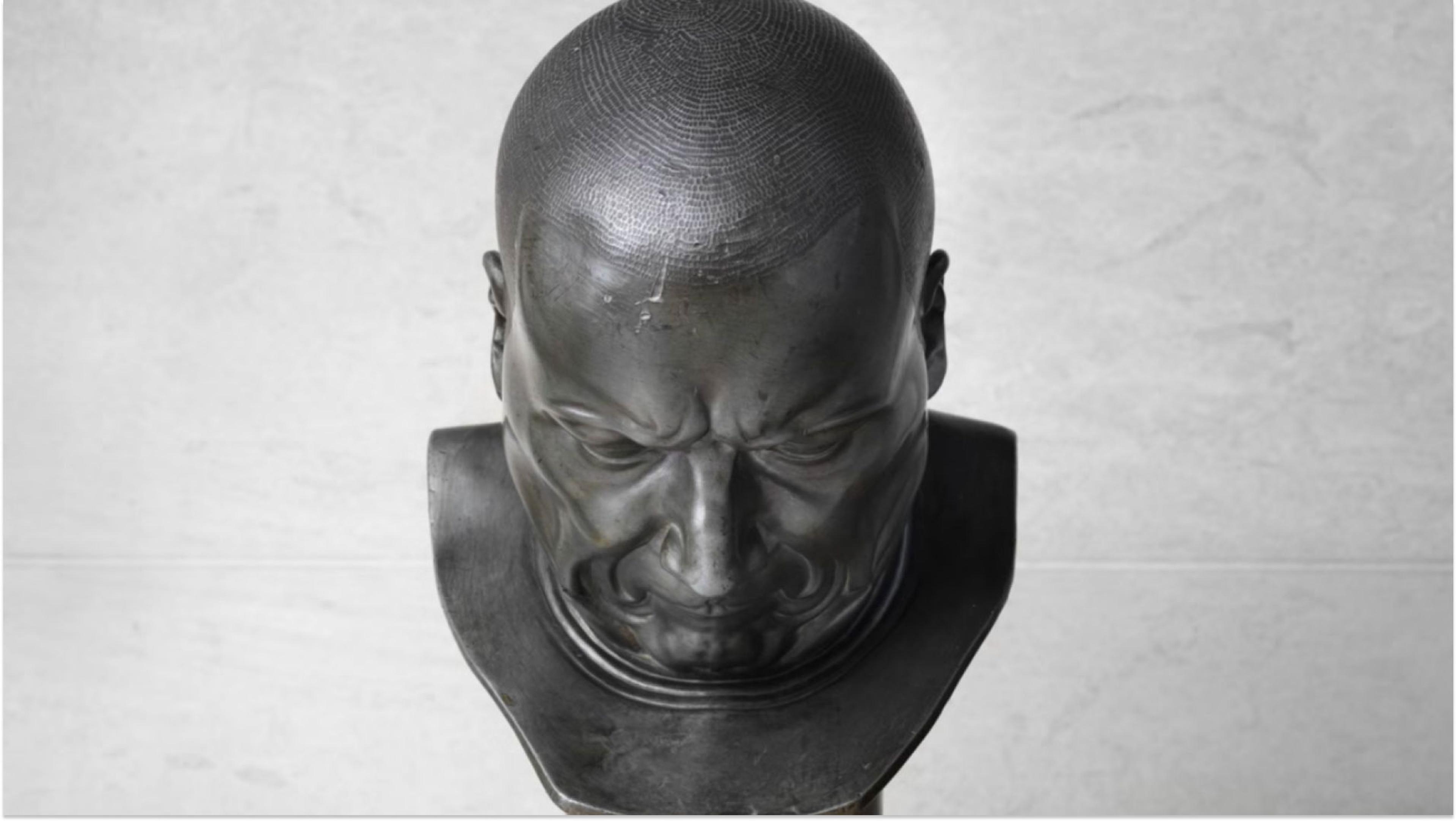 Metal bust with a head is looking down with a concerned expression, a wrinkled brow, and tension in the face. Behind is a white-grey background. 