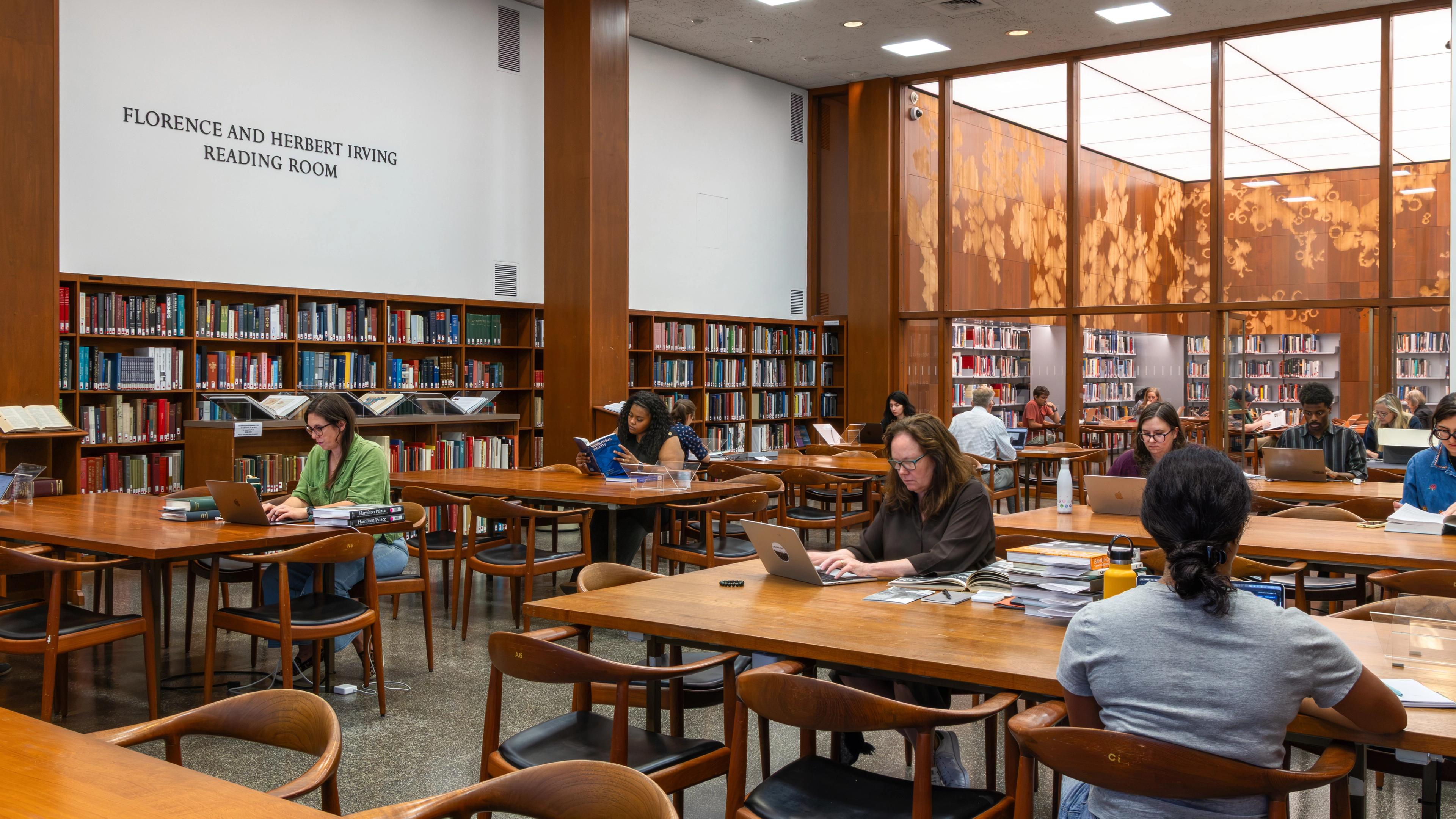 Reading room with bookshelves and study tables; visitors read and work on laptops beneath a sign reading “Florence and Herbert Irving Reading Room.”