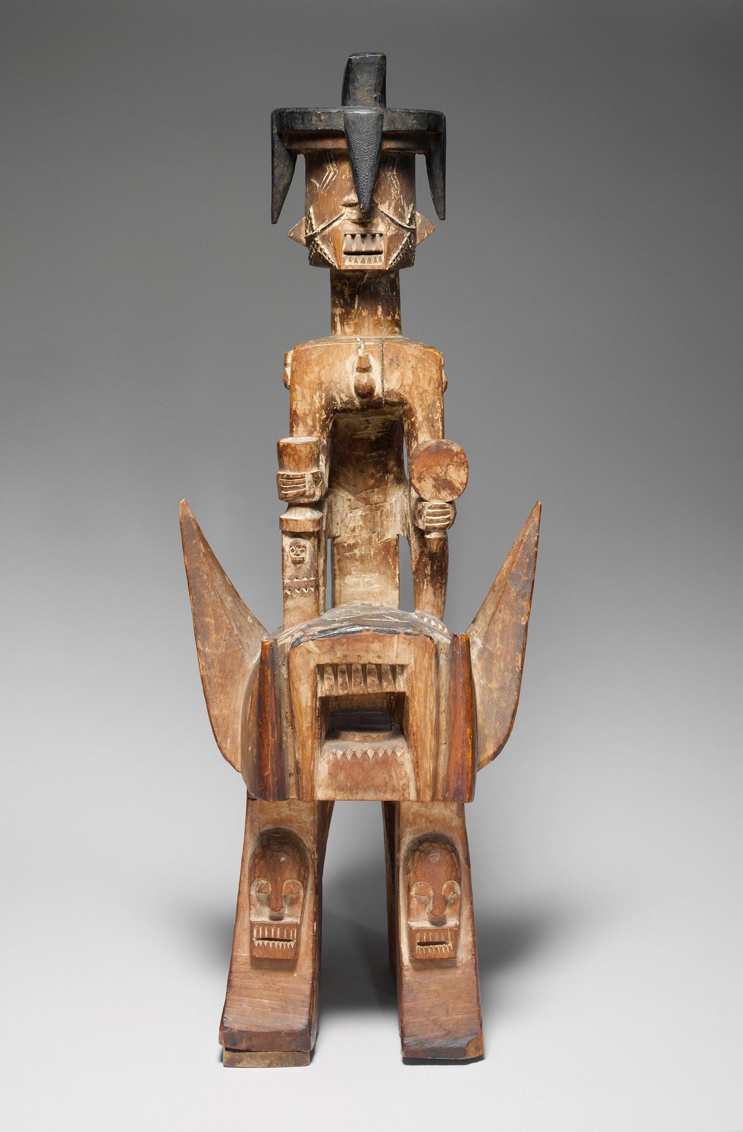 Wooden sculpture of a stylized figure with a geometric face, seated on an abstract horse with an open-mouthed head.
