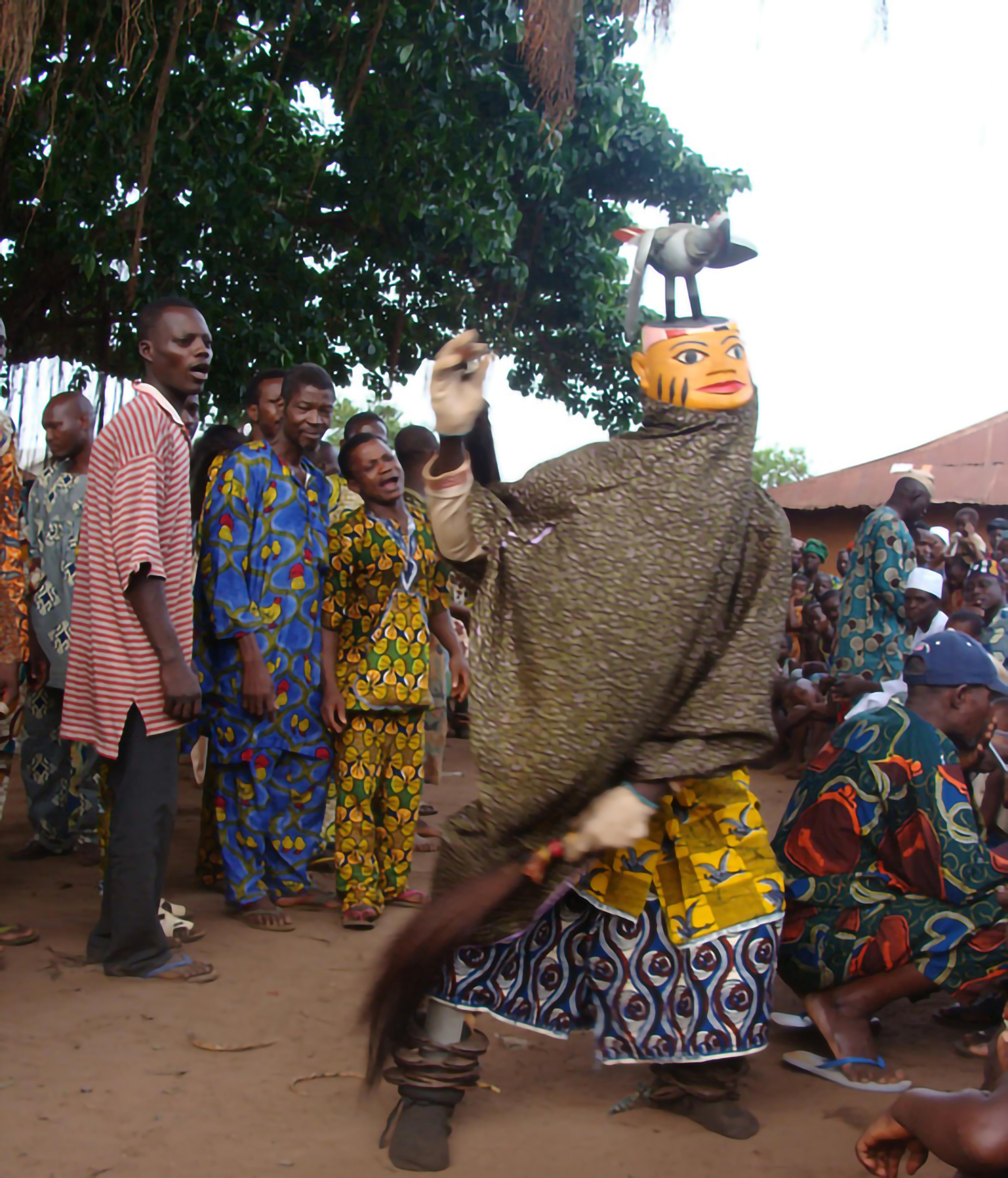 An outdoor scene. The central figure wears a mask topped by a bird while onlookers watch.