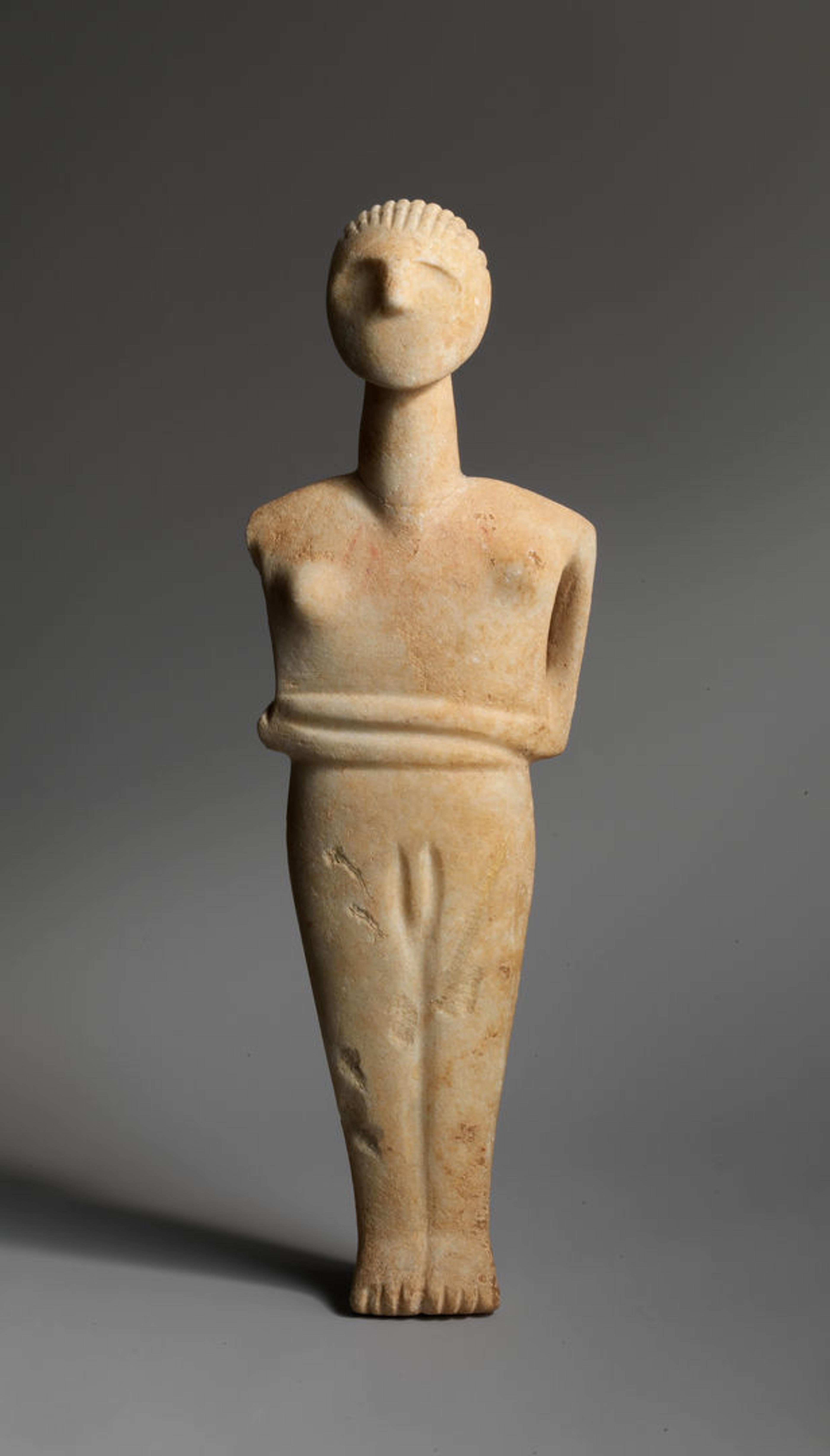 Nude stone Cycladic figure with it's arms crossed against a grey background.