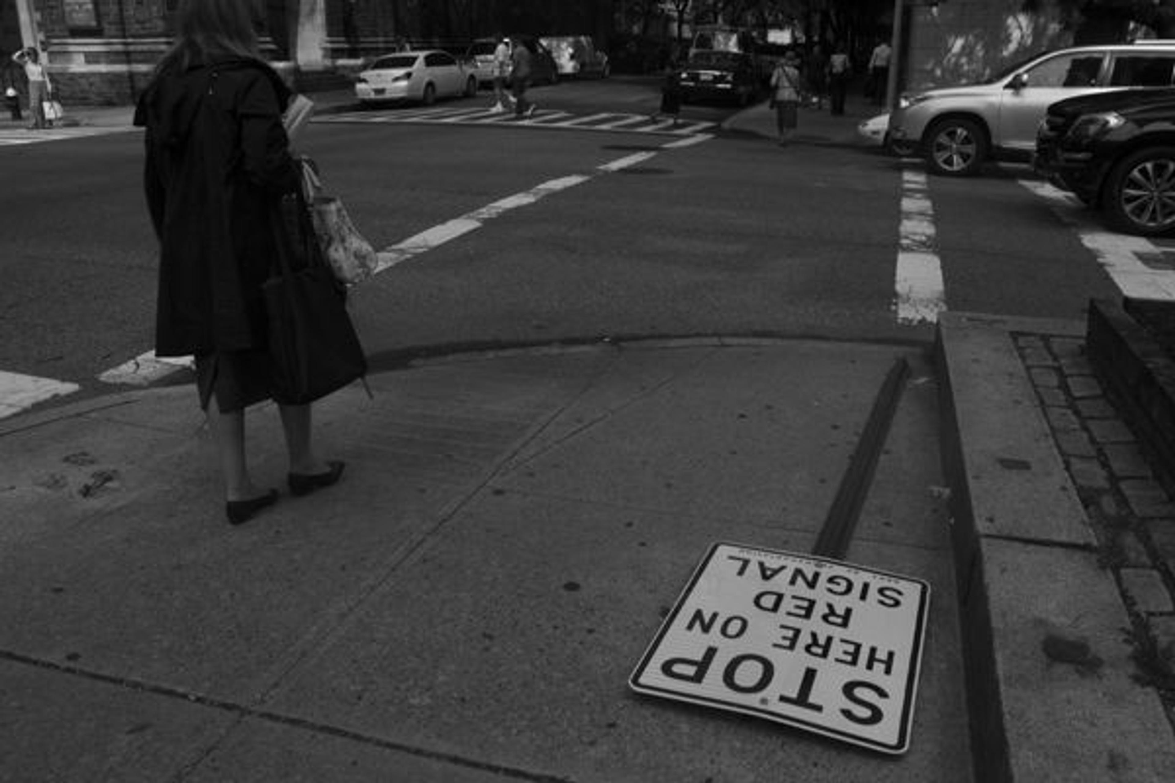 Woman crossing the street next to a fallen sign reading "Stop Here on Red Signal"