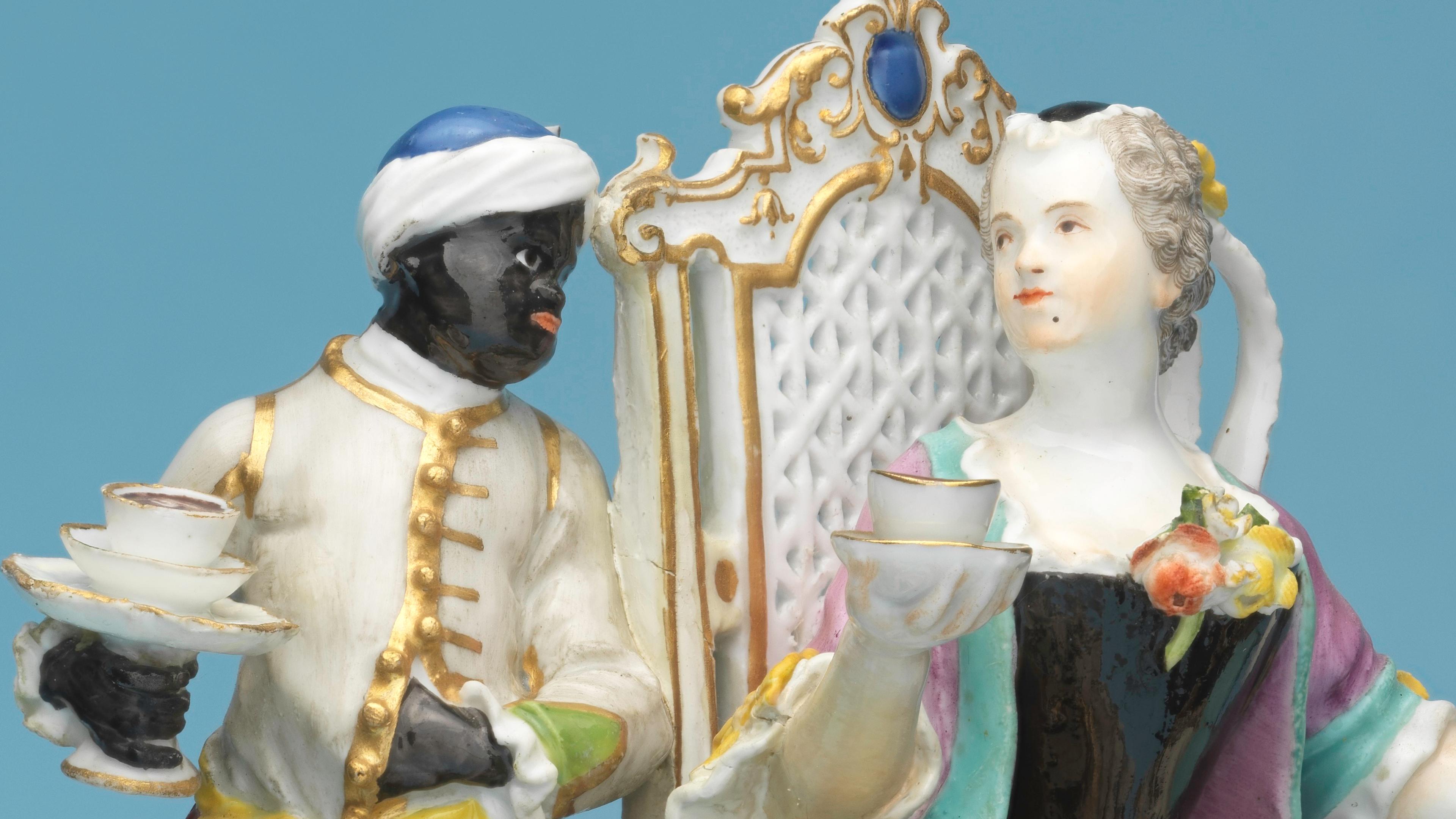 Porcelain figurine depicting an 18th-century scene with a servant holding a tray of cups and a seated aristocratic woman in floral attire, against a blue background.