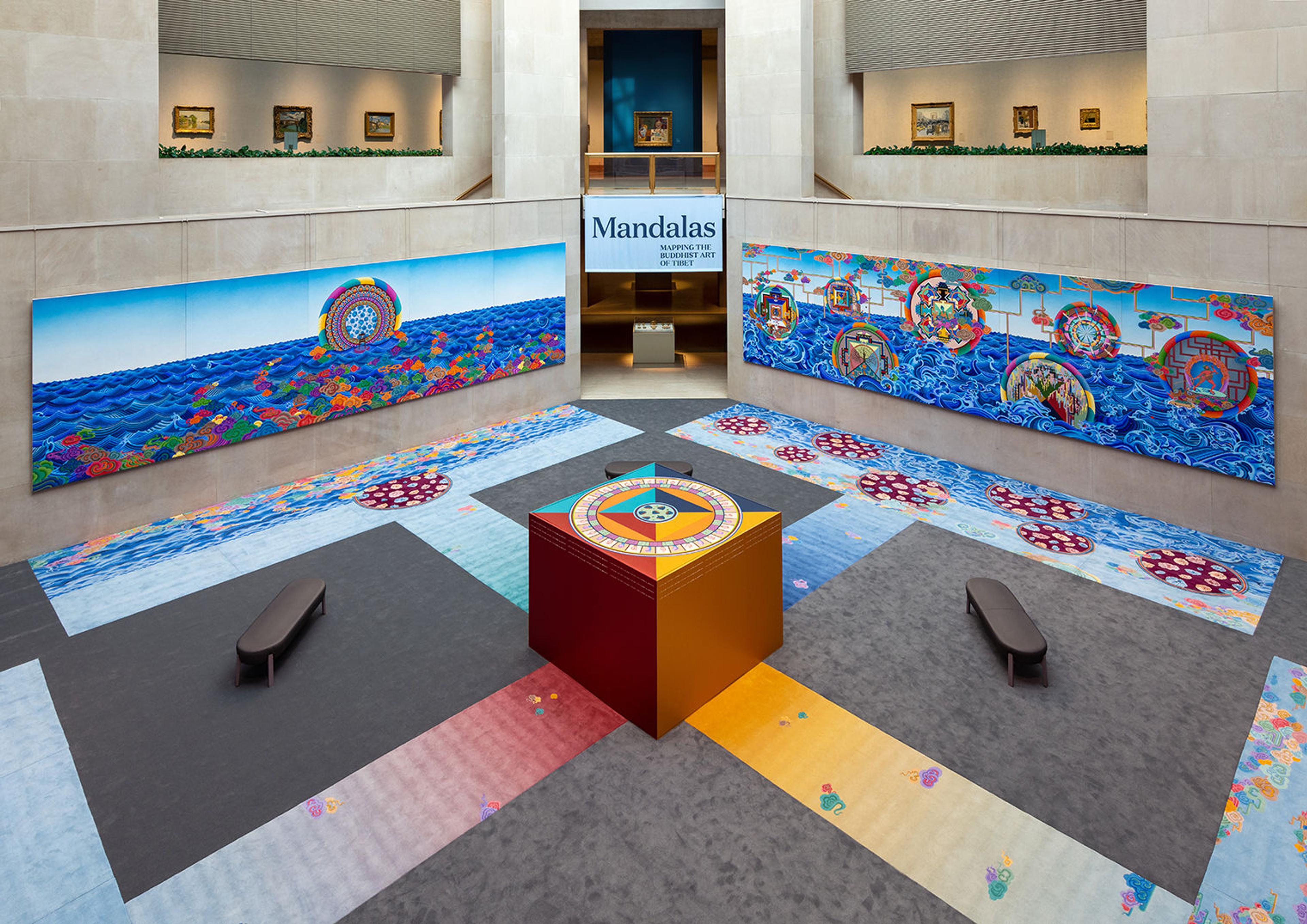 Shot of two large paintings, carpeted illustrations, and a large painted box with a Mandala on top of it in a large, skylit gallery.
