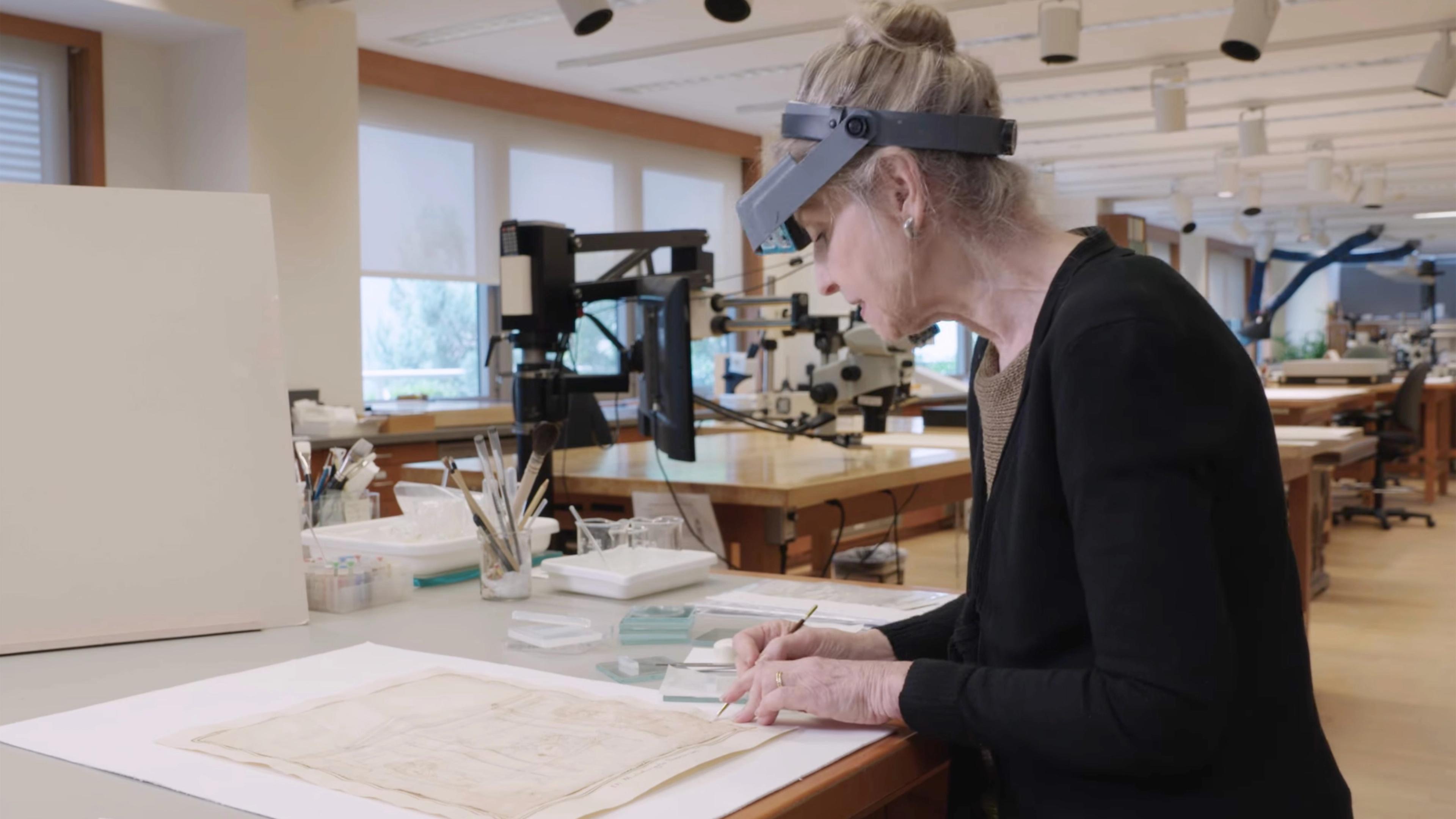 A conservator wearing goggles stands over a drawing on a table