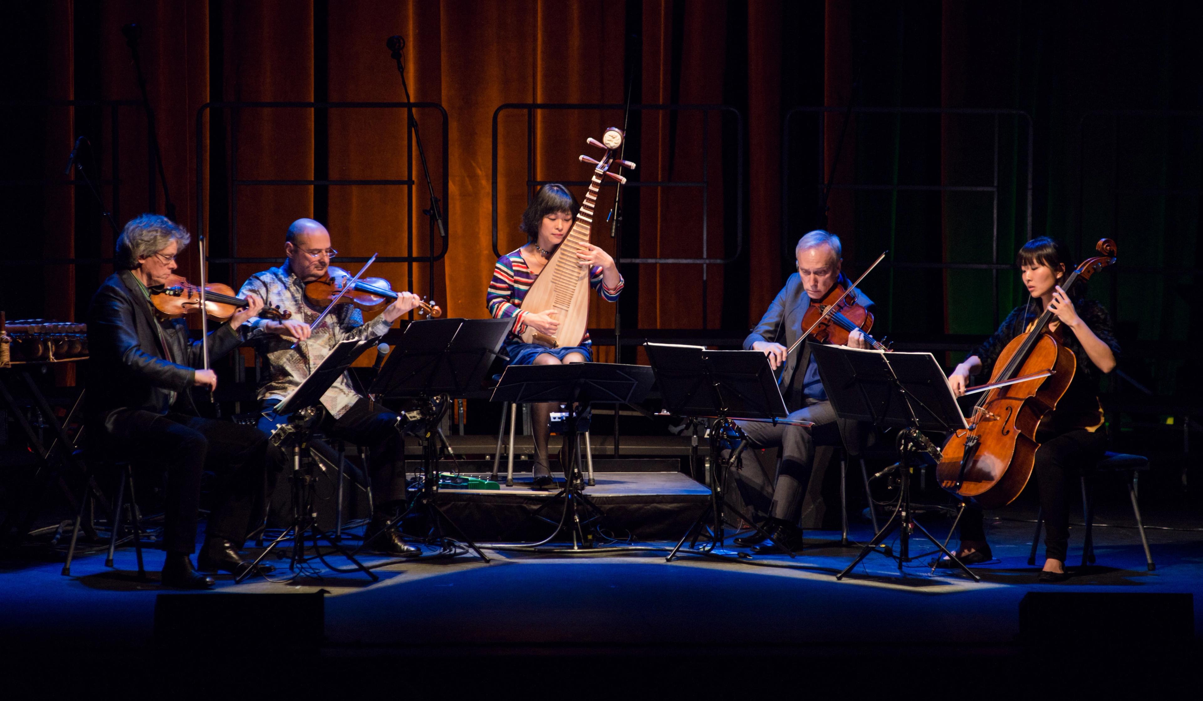 Wu Man at center playing the pipa, flanked by two musicians on either side, on a dark stage.