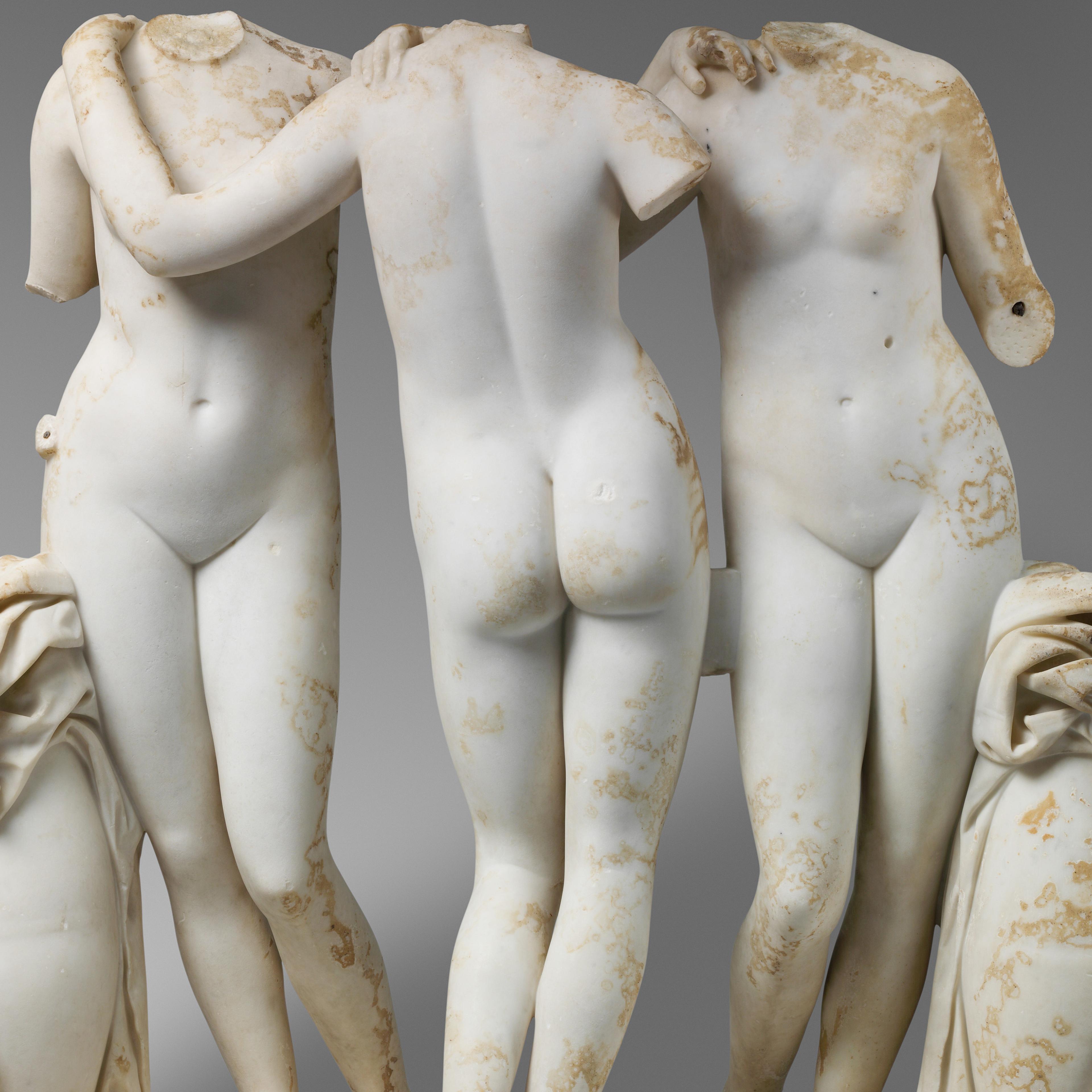 Marble statue of three young women standing in contrapposto between two urns, with fabric on top, while holding on to each other's shoulders