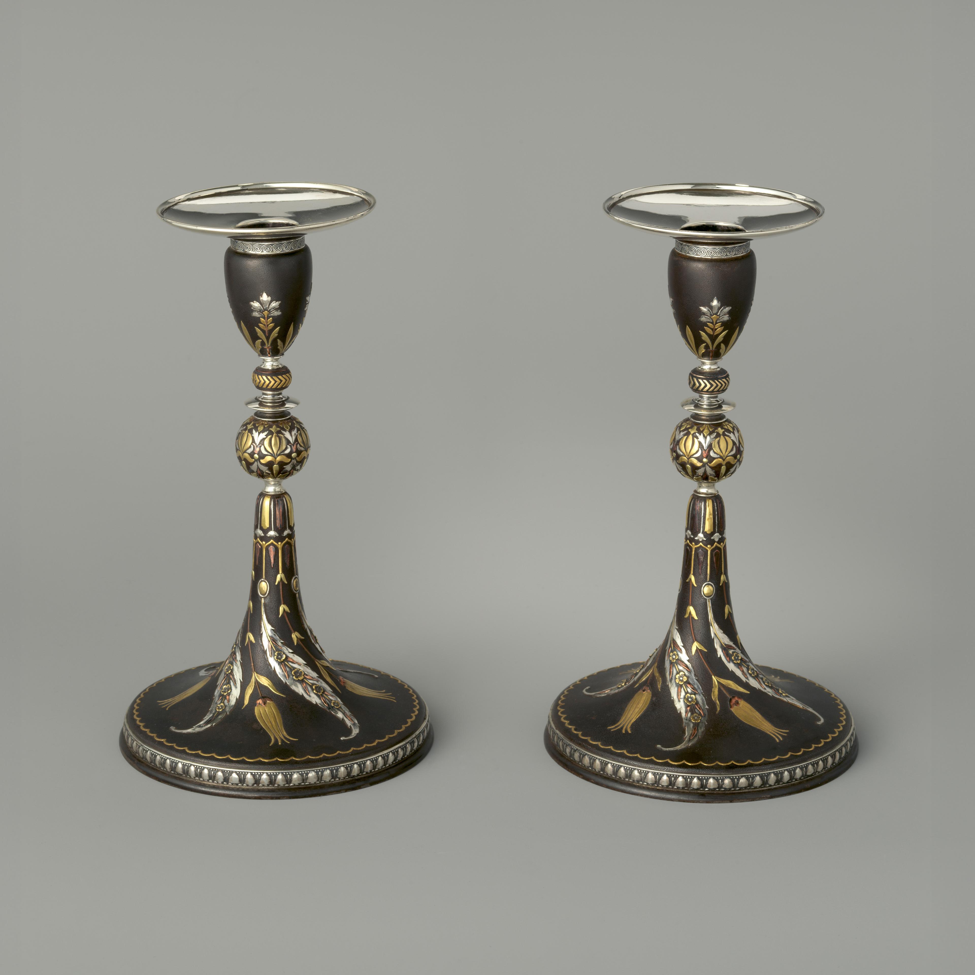 A pair of black iron candlesticks with a tapered conical base appear side-by-side. They are adorned with various sinuous floral motives that are modeled in low relief on the surface and surfaced in a variety of silver-, gold-, and copper-toned metals.