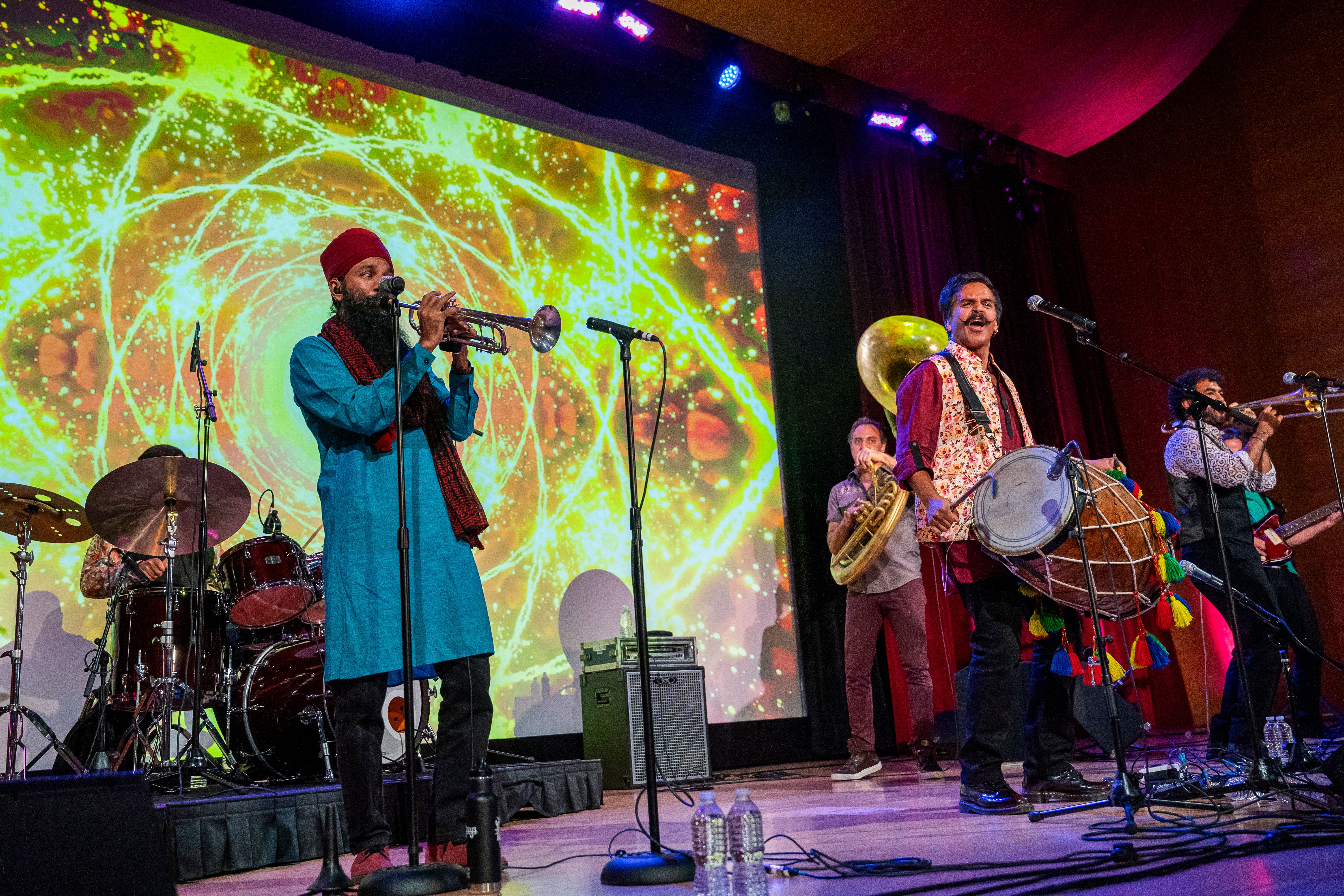 A drummer, a trumpet player, a dhol player, a sousaphone player, and a trombone player perform on a stage in front of psychedelic projections.