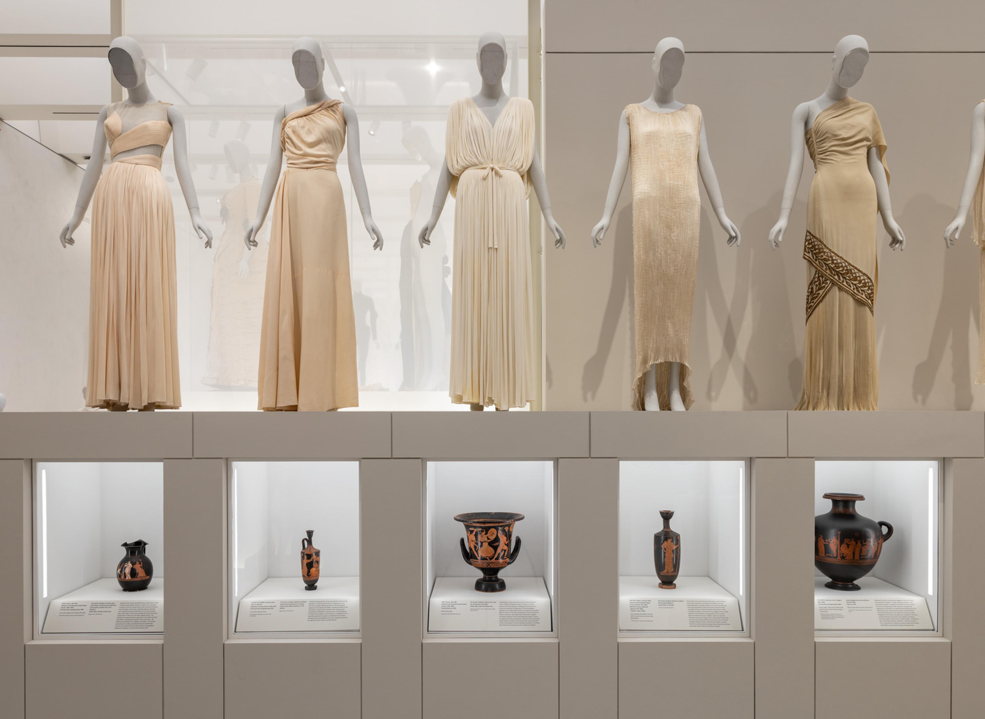 Five off-white gowns with similar columnar shapes dressed on gray mannequins, each paired with a Classical ceramic vessel painted with people wearing similar garments.