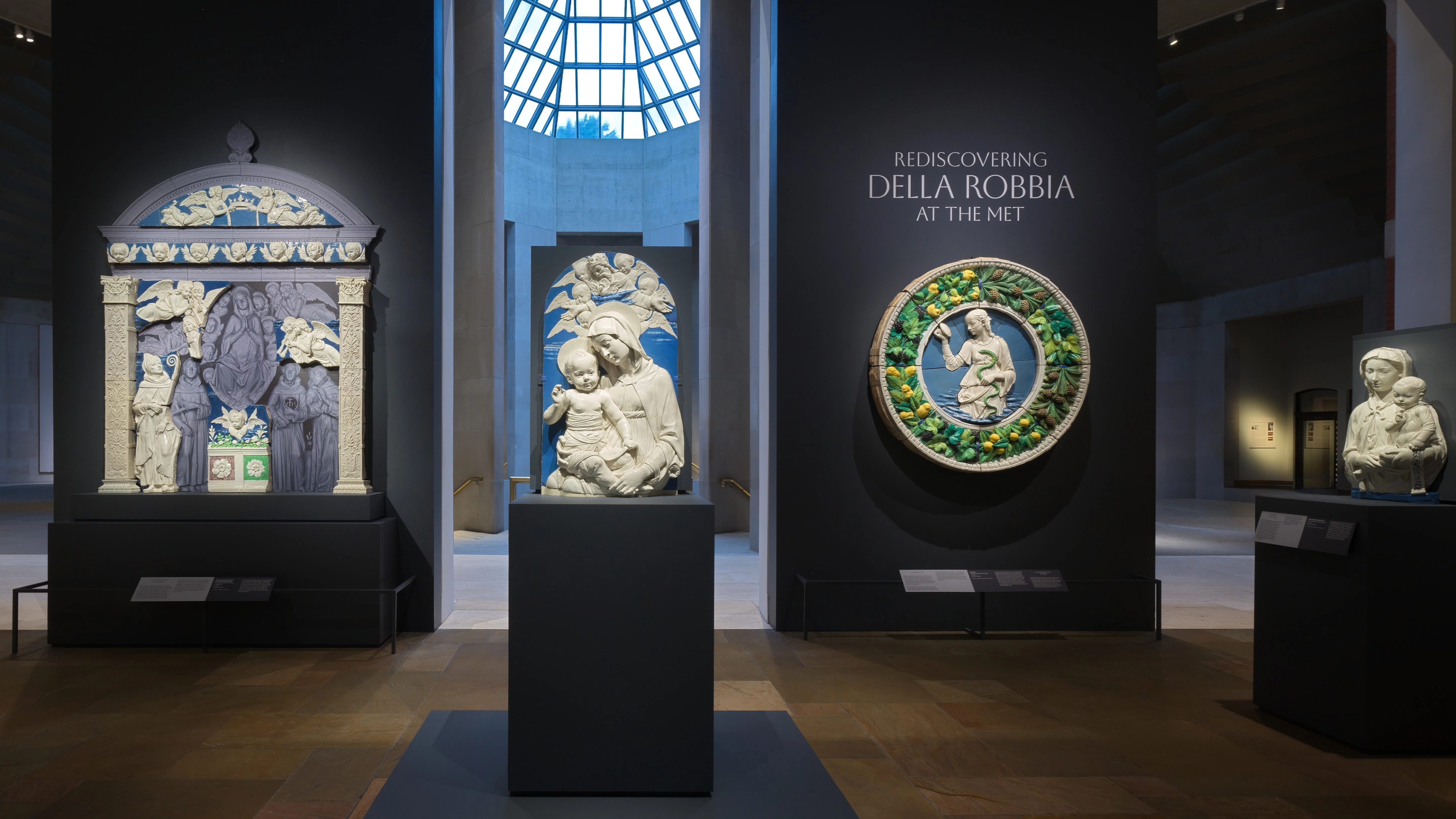 A quiet museum gallery displays Della Robbia ceramics: a colorful altar panel, a white sculpted Madonna and child, and a green circular relief.