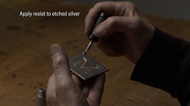 An artisan uses a small brush to paint a resist material on the sides of a small metal plate