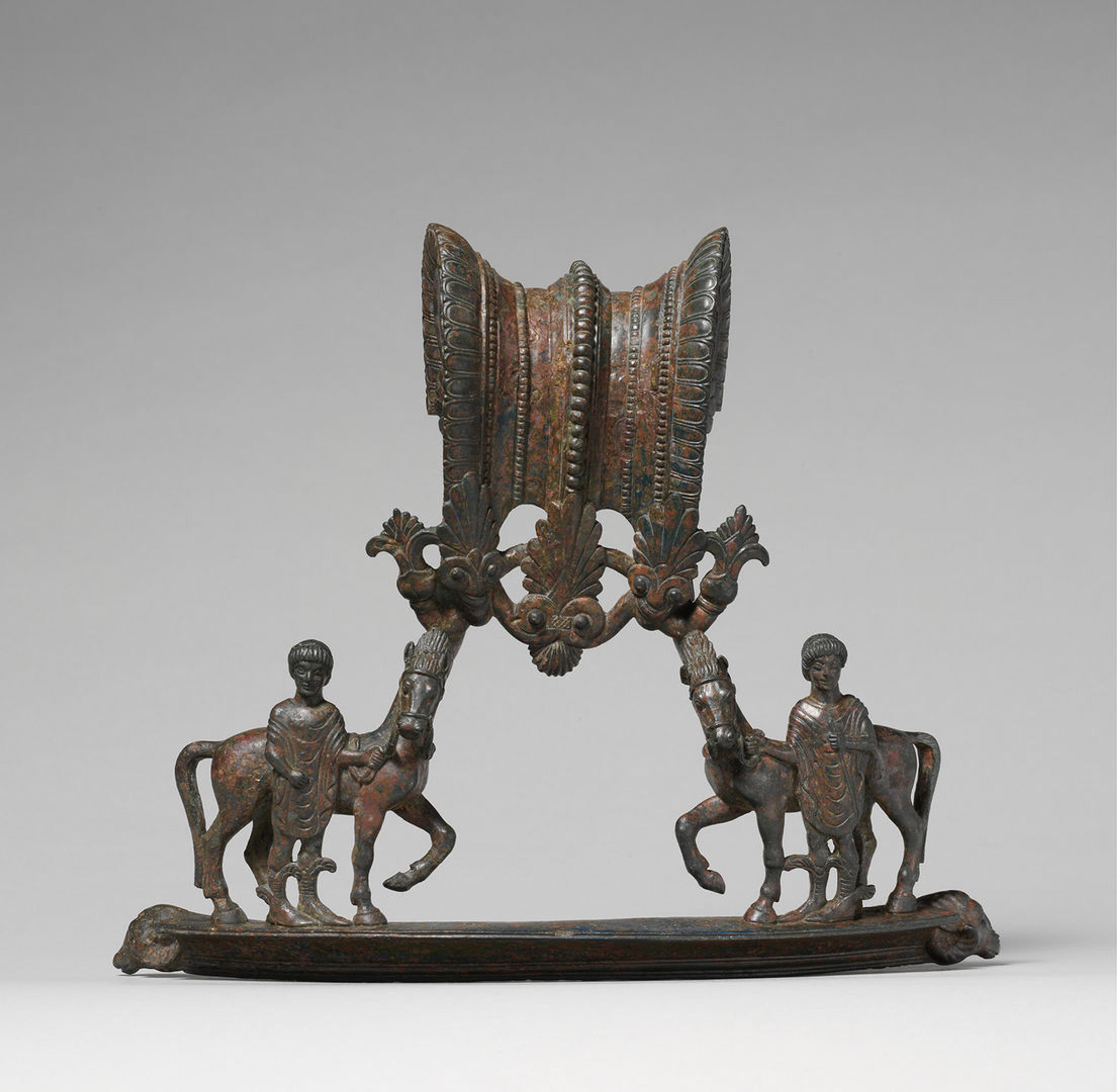 Bronze handles from a volute-krater depicting two youths with winged boots, holding the bridles of their horses. Elaborately worked bronze links the two figures above the horses' heads.