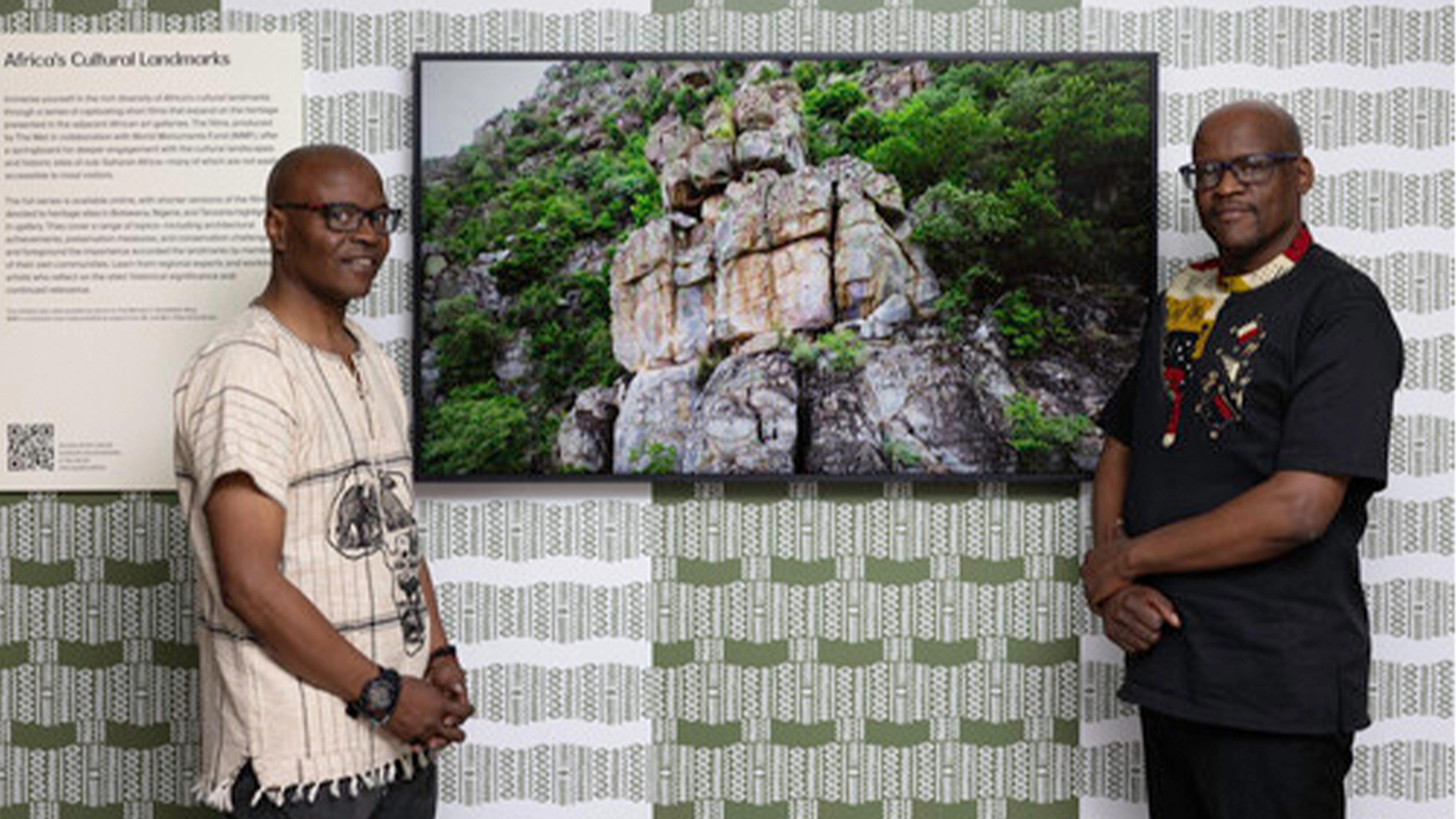 Two individuals stand beside a television displaying a vibrant photograph of rock formations amidst greenery, against a patterned backdrop.