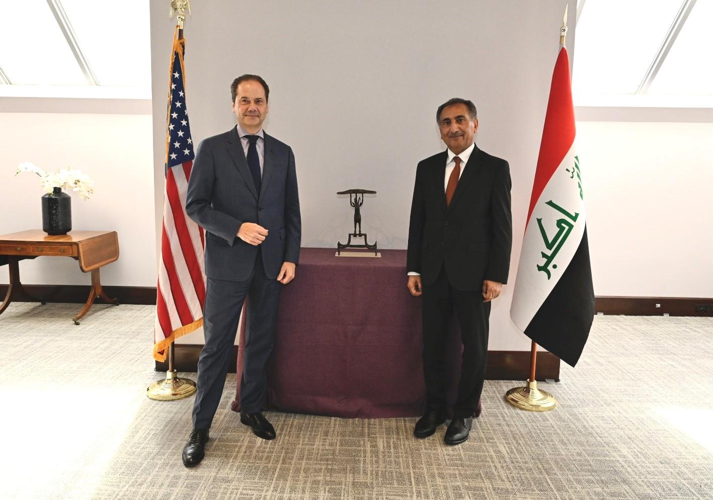 Two men in suits standing in front of a bronze sculpture flanked by an American and Iraqi flag