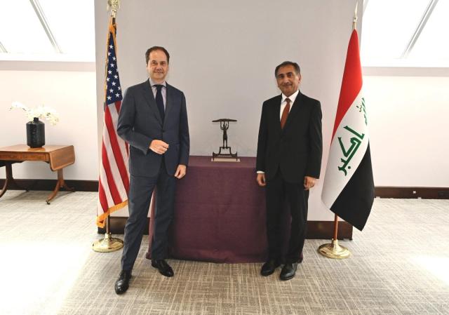 Two men in suits standing in front of a bronze sculpture flanked by an American and Iraqi flag