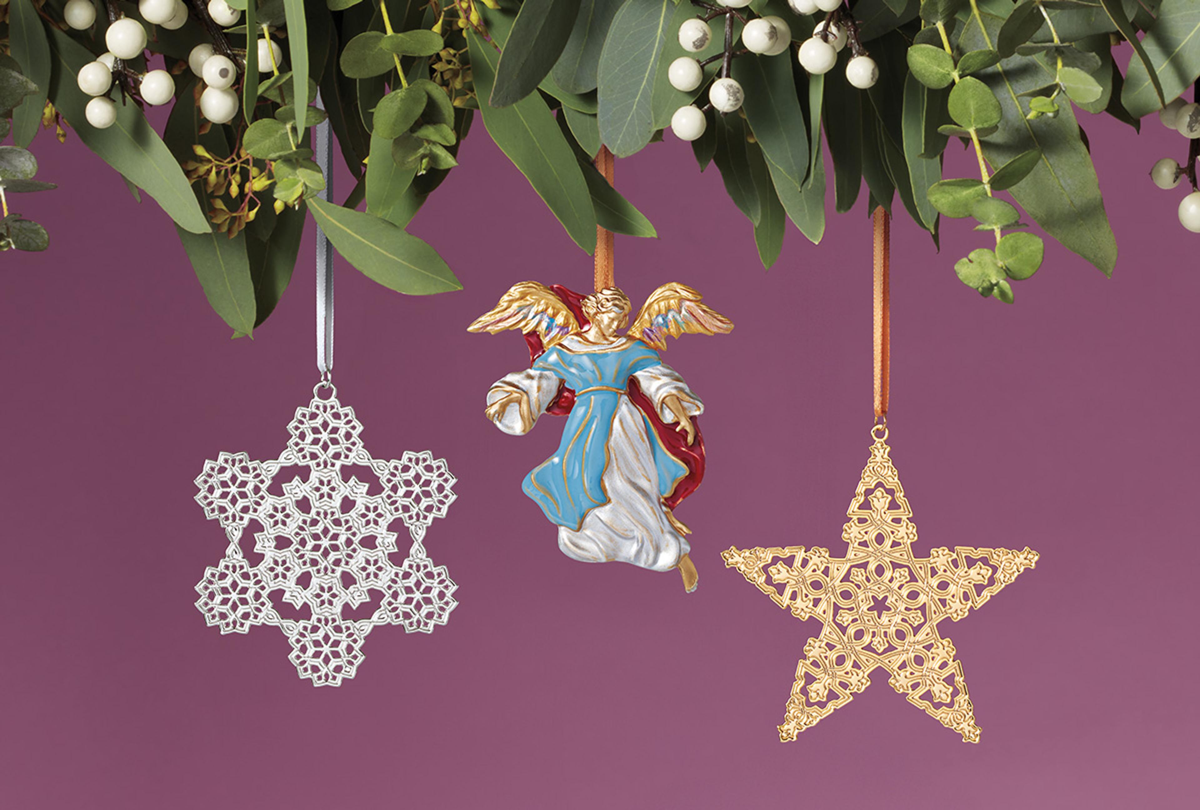 Three holiday ornaments in the shape of a snowflake, angel, and star, hang in front of a violet colored background.