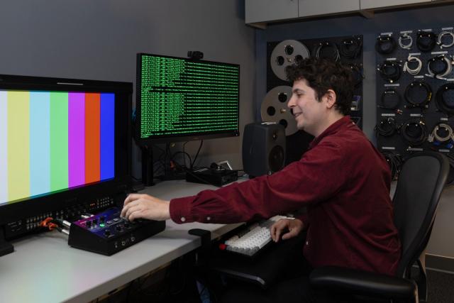 A person sits at a workstation adjusting equipment, with a color bars test pattern on one monitor and code on another, in a room with cables and film reels on the wall.