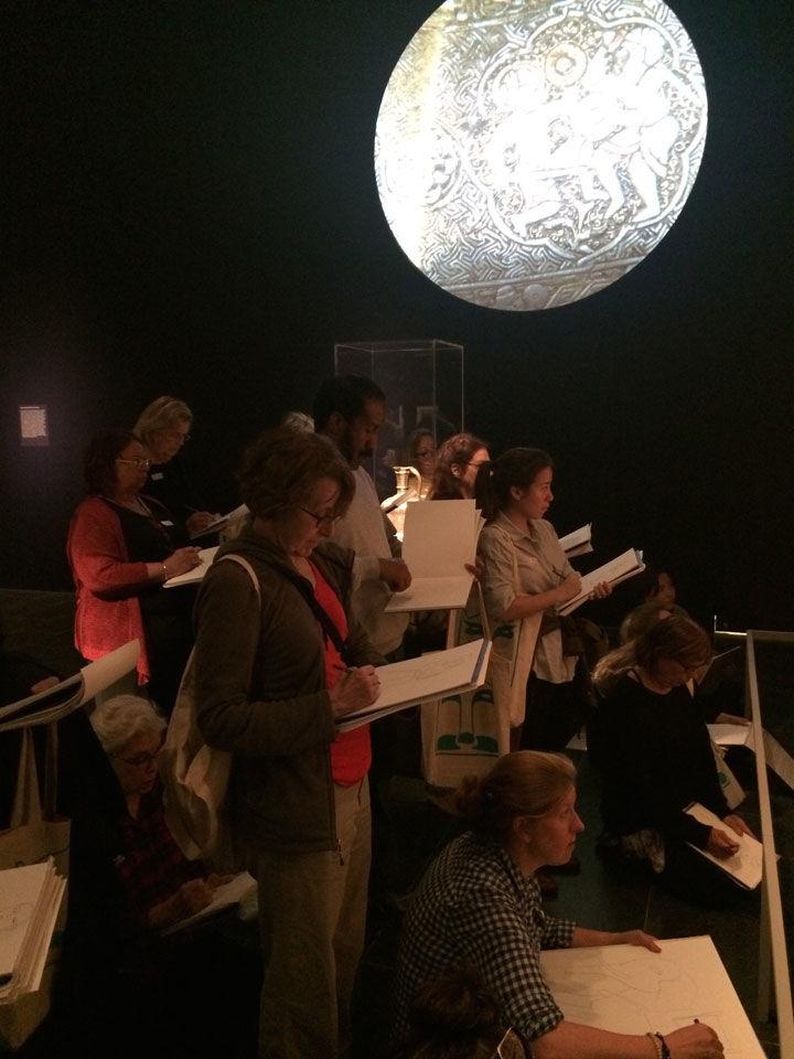 A group of people sketch in a dark exhibition gallery lit by a moon-like object on the wall