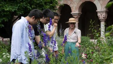 A group of three young people listens attentively to a woman guiding them through a lush garden filled with tall purple flowers and greenery.