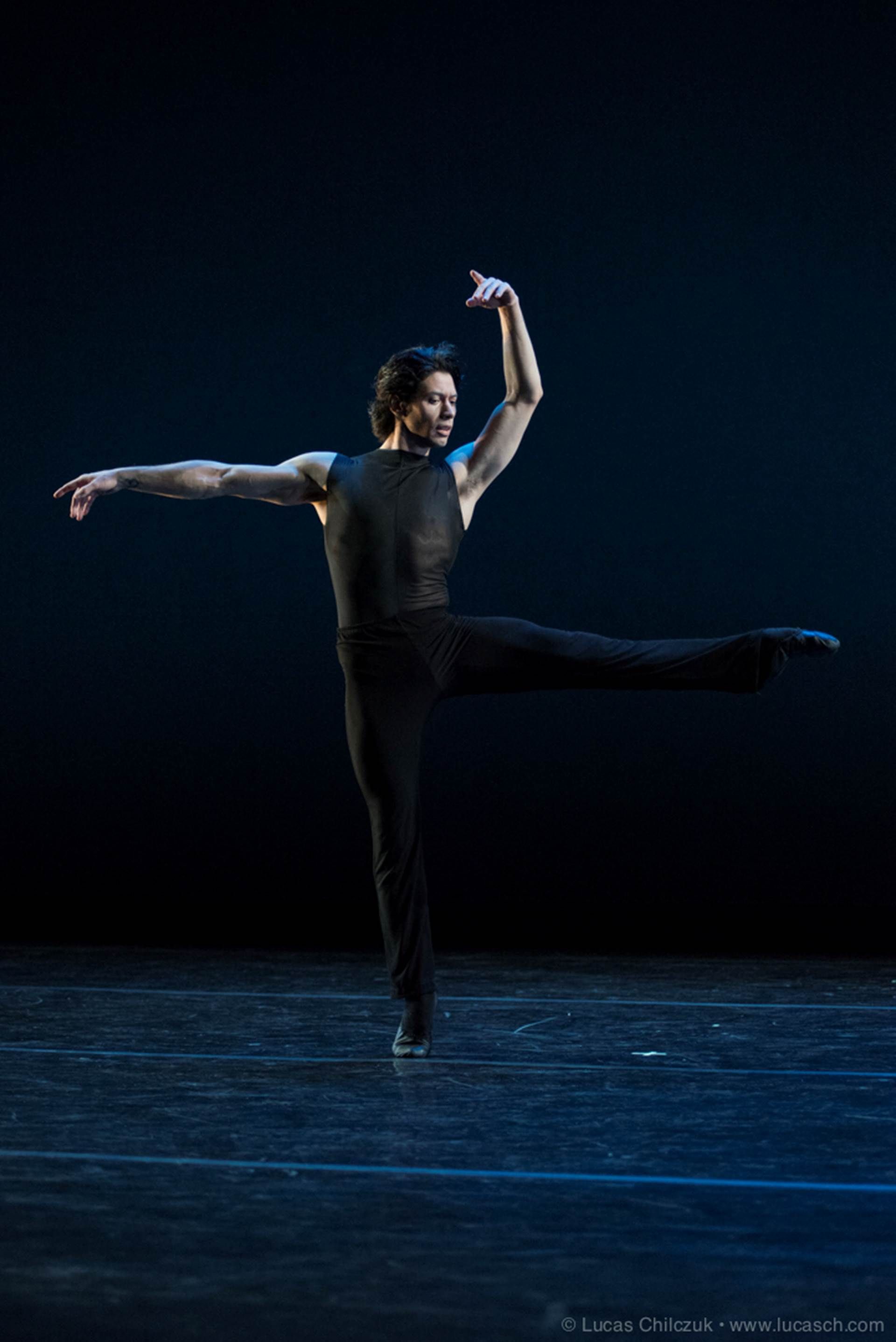 A medium-skin-toned male dancer is photographed mid-chorography in a dramatic dance pose in front of a blackish-blue background.