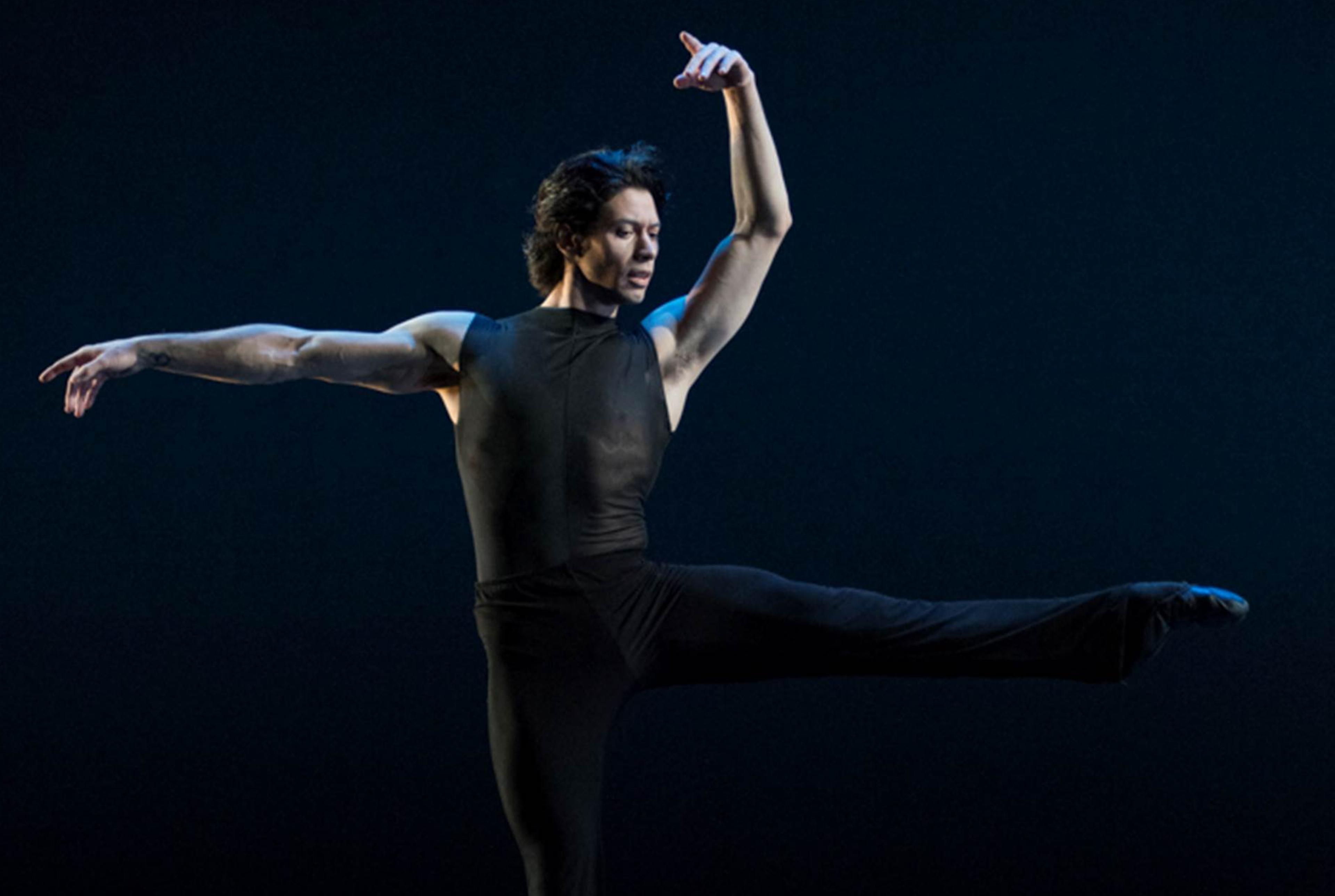 A medium-skin-toned male dancer is photographed mid-chorography in a dramatic dance pose in front of a blackish-blue background.