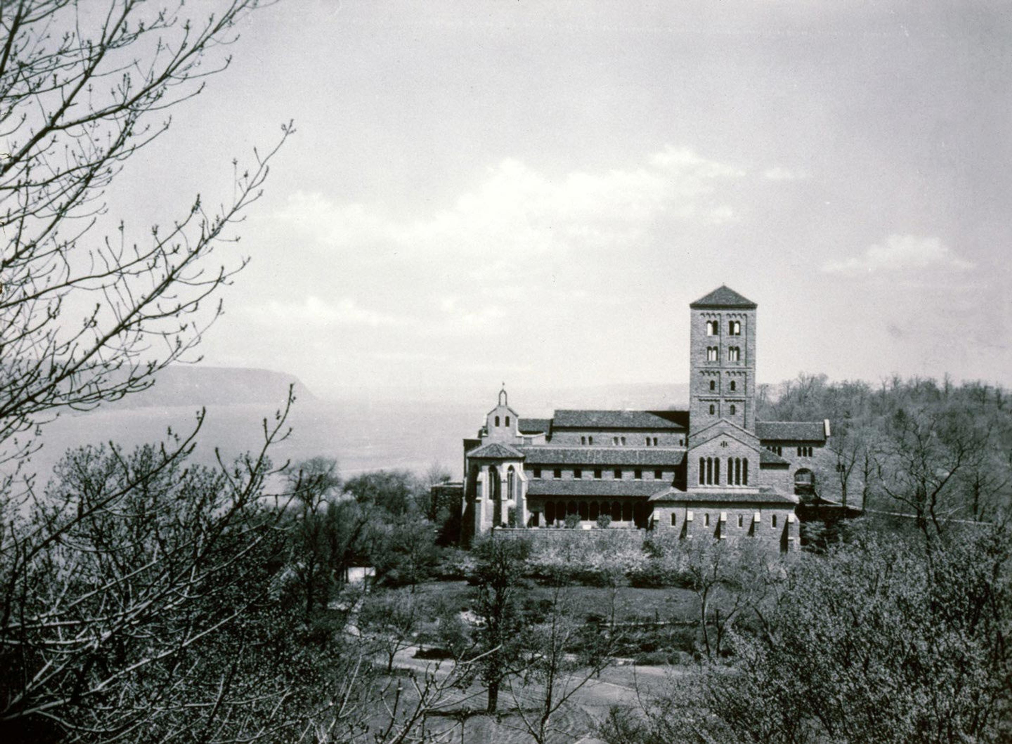 A medieval stone building with a tall tower on the right in a forest landscape, of trees, brush, and a cloudy sky. The large river is visible behind the building in this in black and white photograph. 
