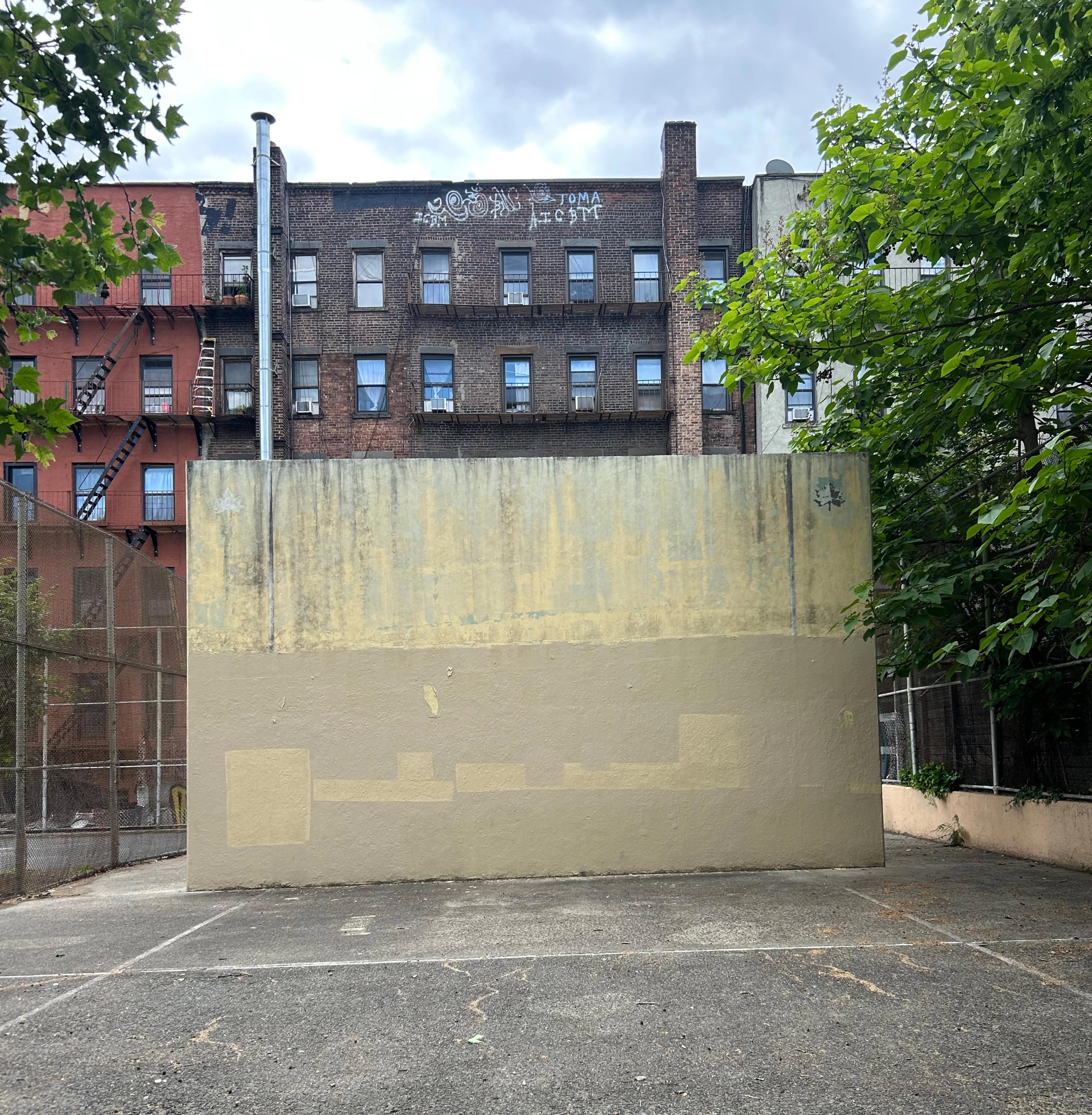 An urban handball court. A row of apartment buildings flank a weather-stained, haphazardly-painted handball wall.