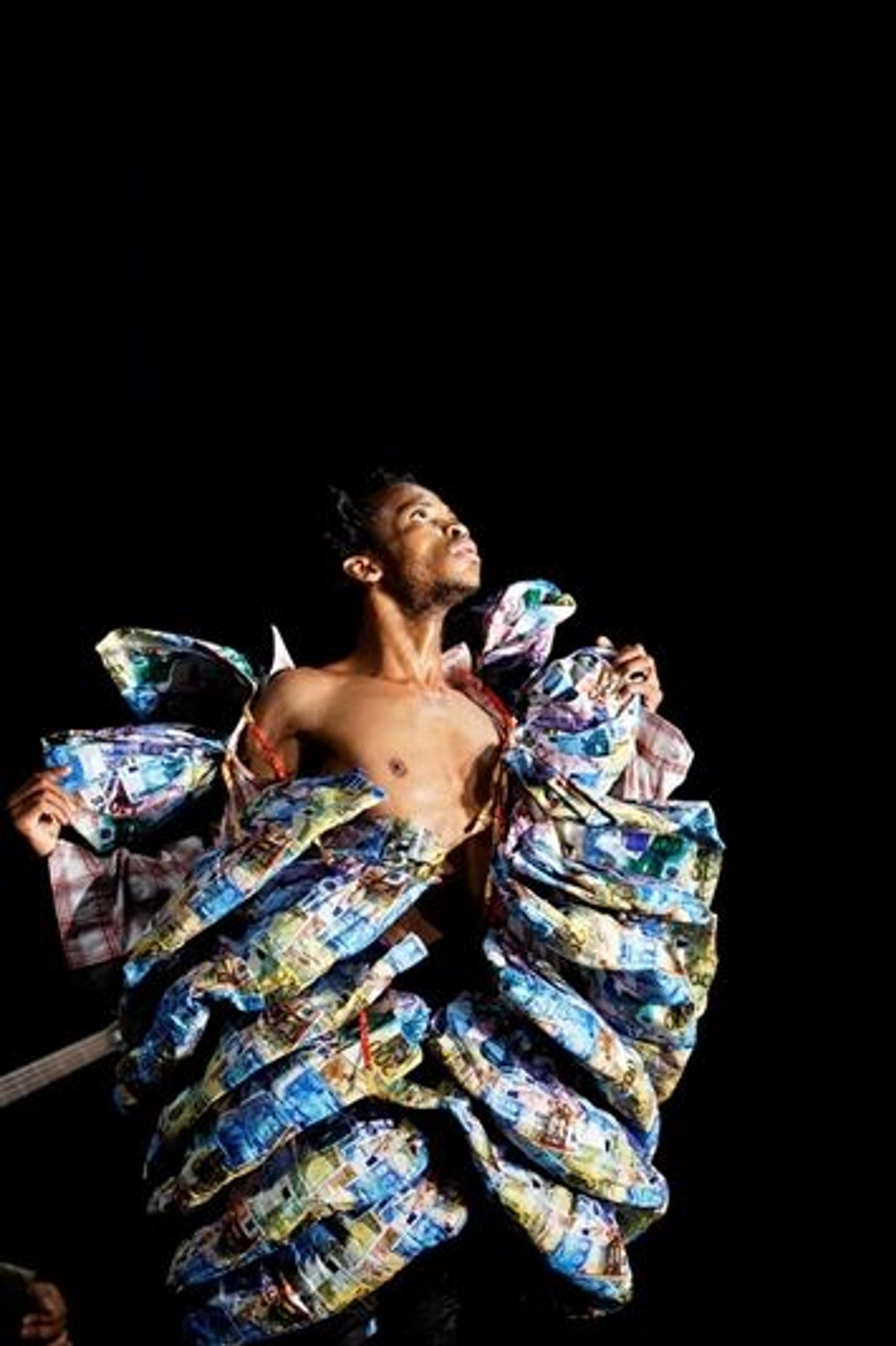 A male dancer against a black background wearing an elaborate costume made of different currencies