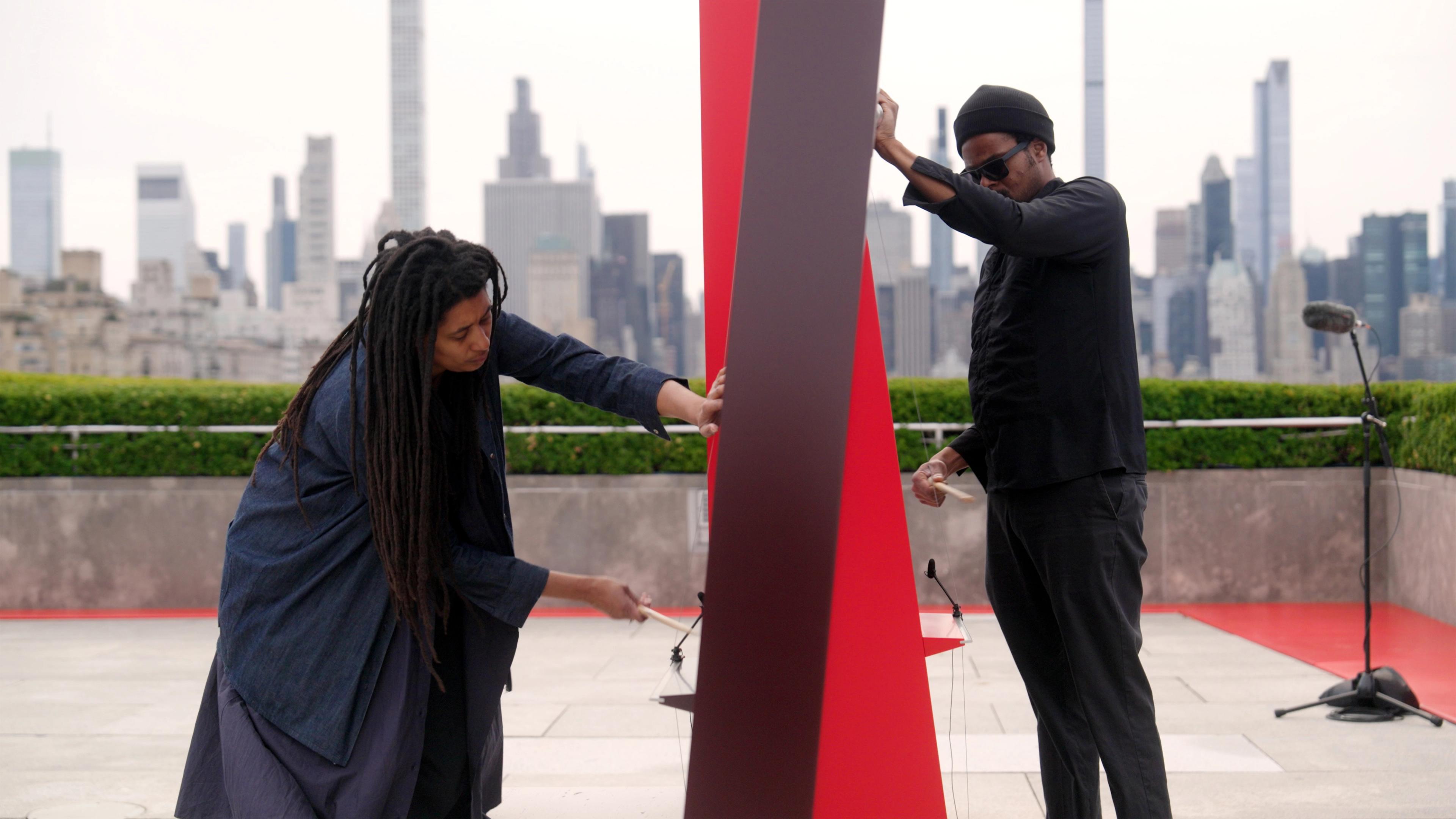 Two individuals interact with a tall, red sculpture on a rooftop terrace. The city skyline is visible in the background, creating an artistic and contemplative atmosphere.