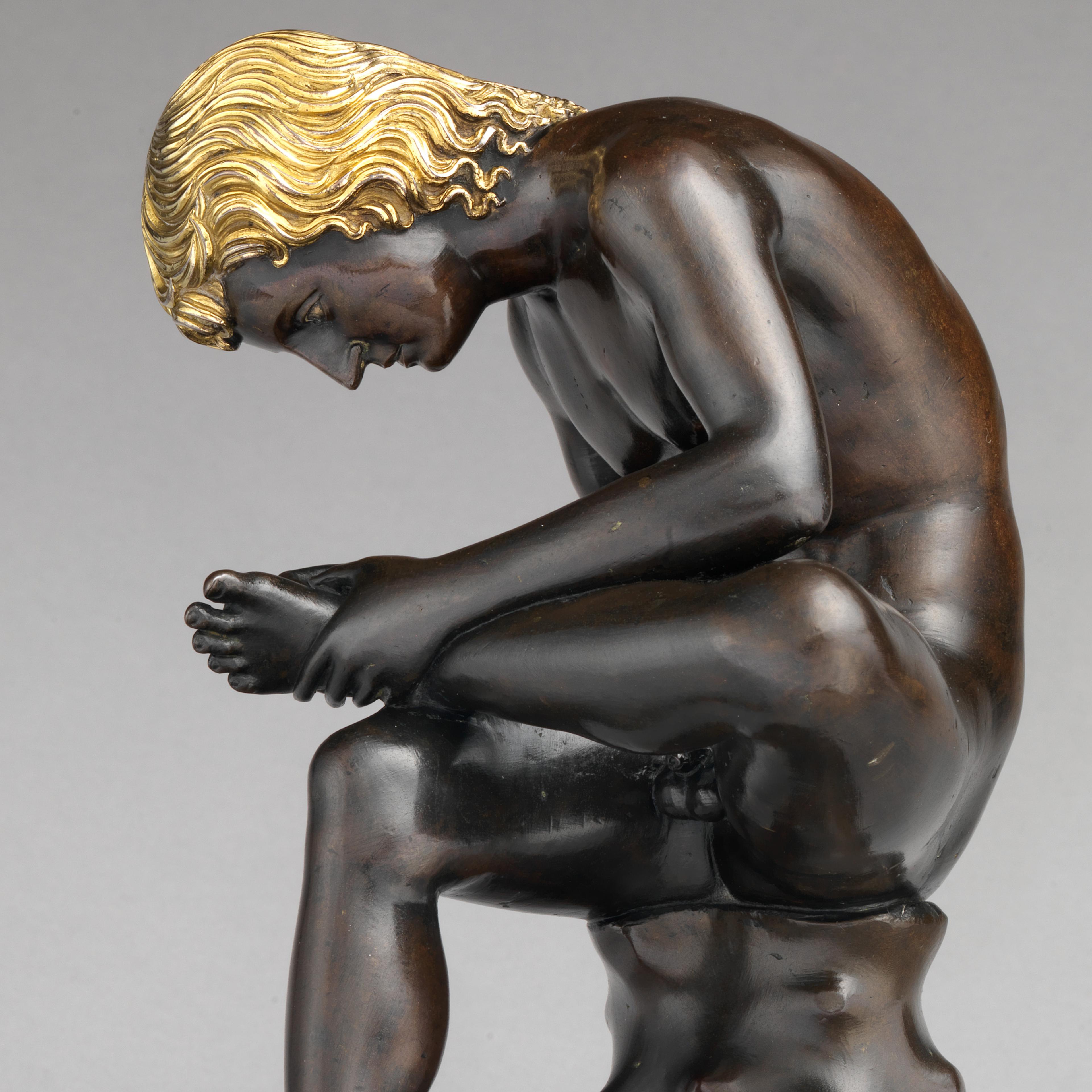 Bronze sculpture of a young boy with gold wavy hair pulling out a thorn from his foot while sitting on a tree stump