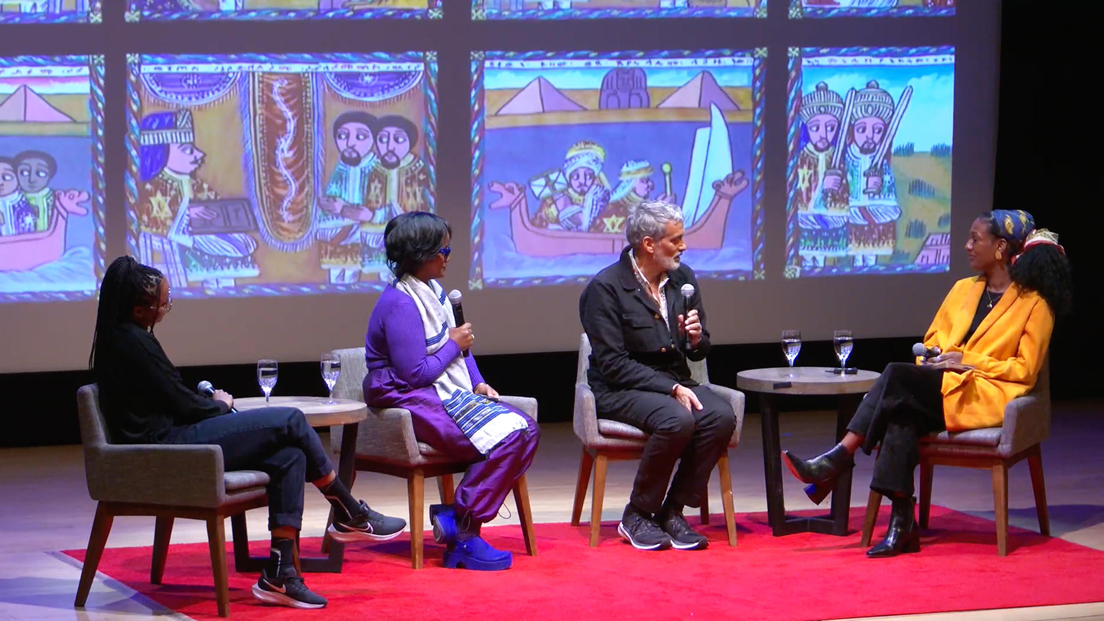 Three artists and one moderator sitting in modern grey chairs on a stage each holdings microphones with a red carpet underneath them. There is a projection screen behind them.