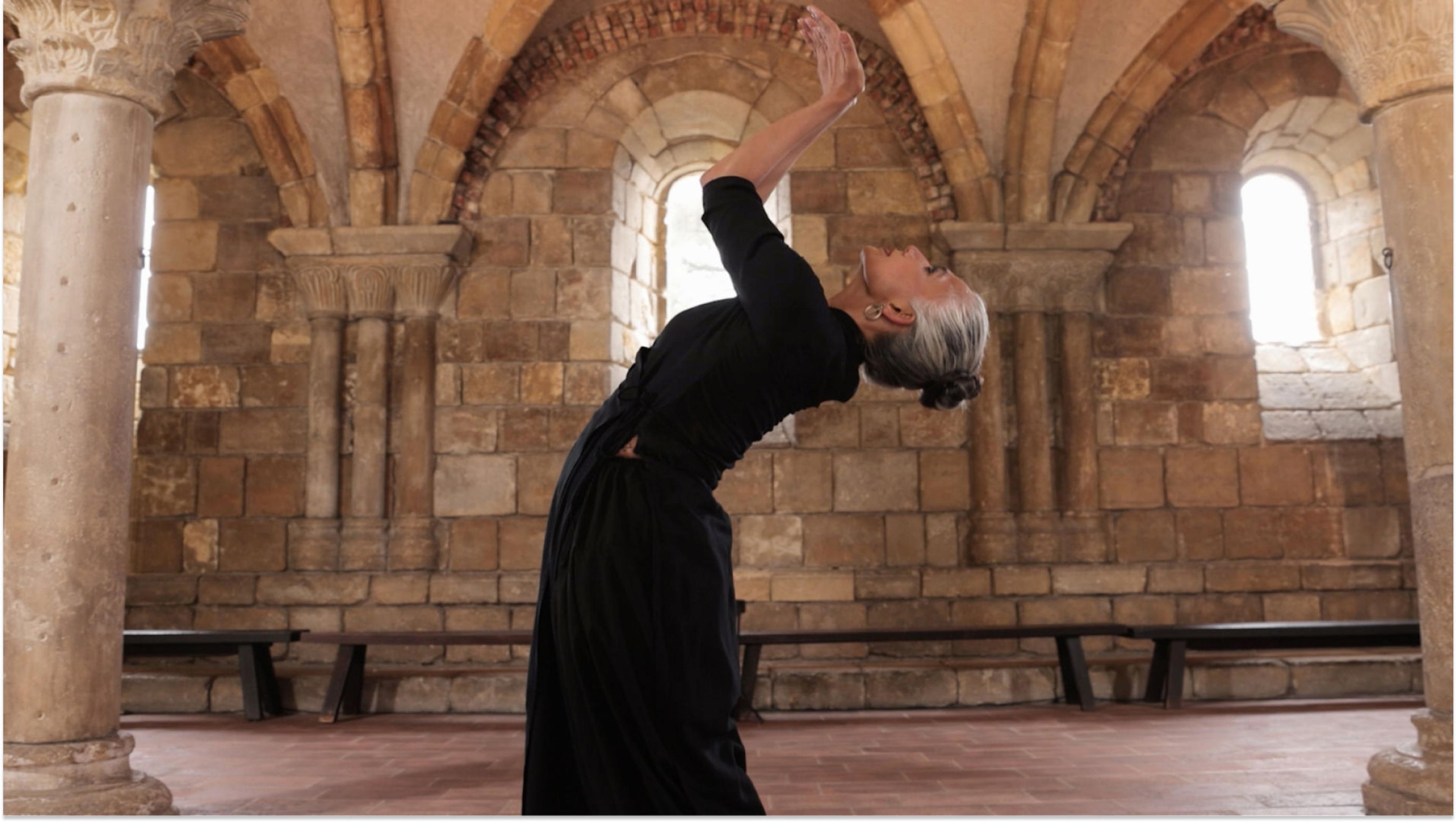 A woman with grey hair and a black dress arches her back, tilts her head back, and raises her hands in a cupped position. She is in a building made of tan stone walls and columns with arched windows.