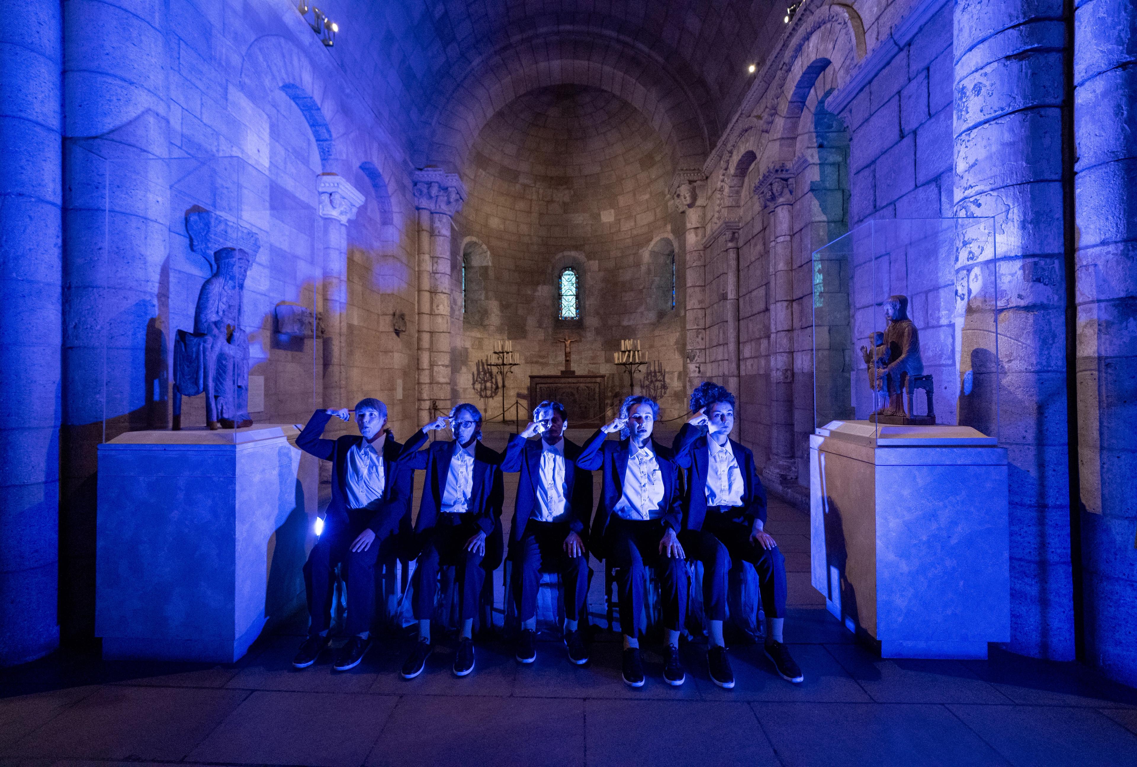 A chapel awash in blue light. In the foreground, five actors sit along a bench, each pointing their index finger against their temples.